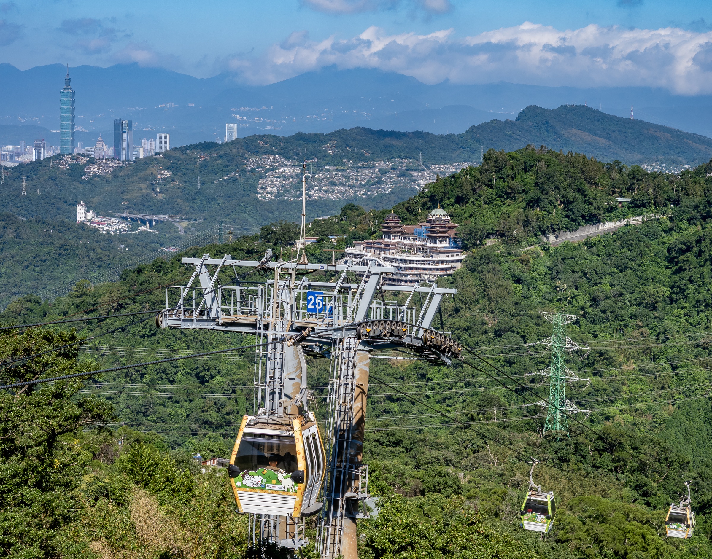 A yellow cable car ascends a steep, green hillside with mountains and city visible in the distance.