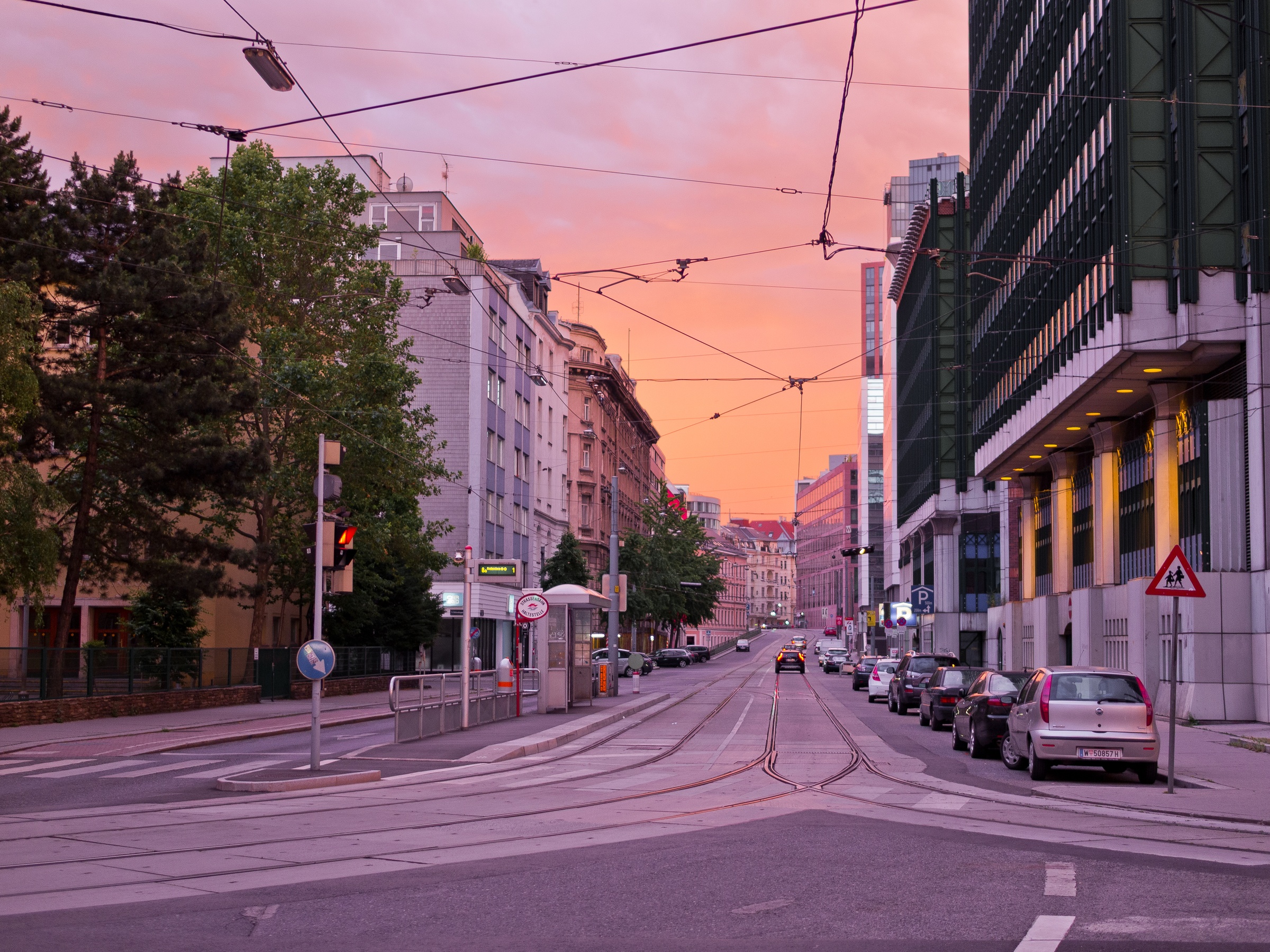 A street with tram tracks and parked cars stretches toward a city skyline under a pink sunset.