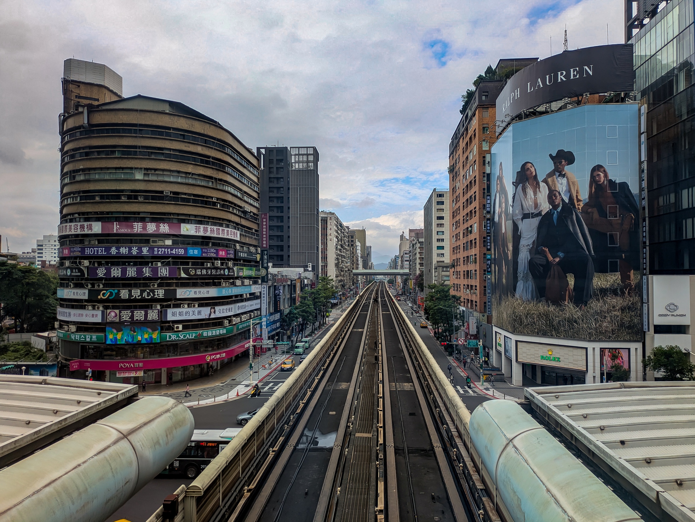 An elevated train track runs between buildings with advertising and a modern cityscape under cloudy skies.