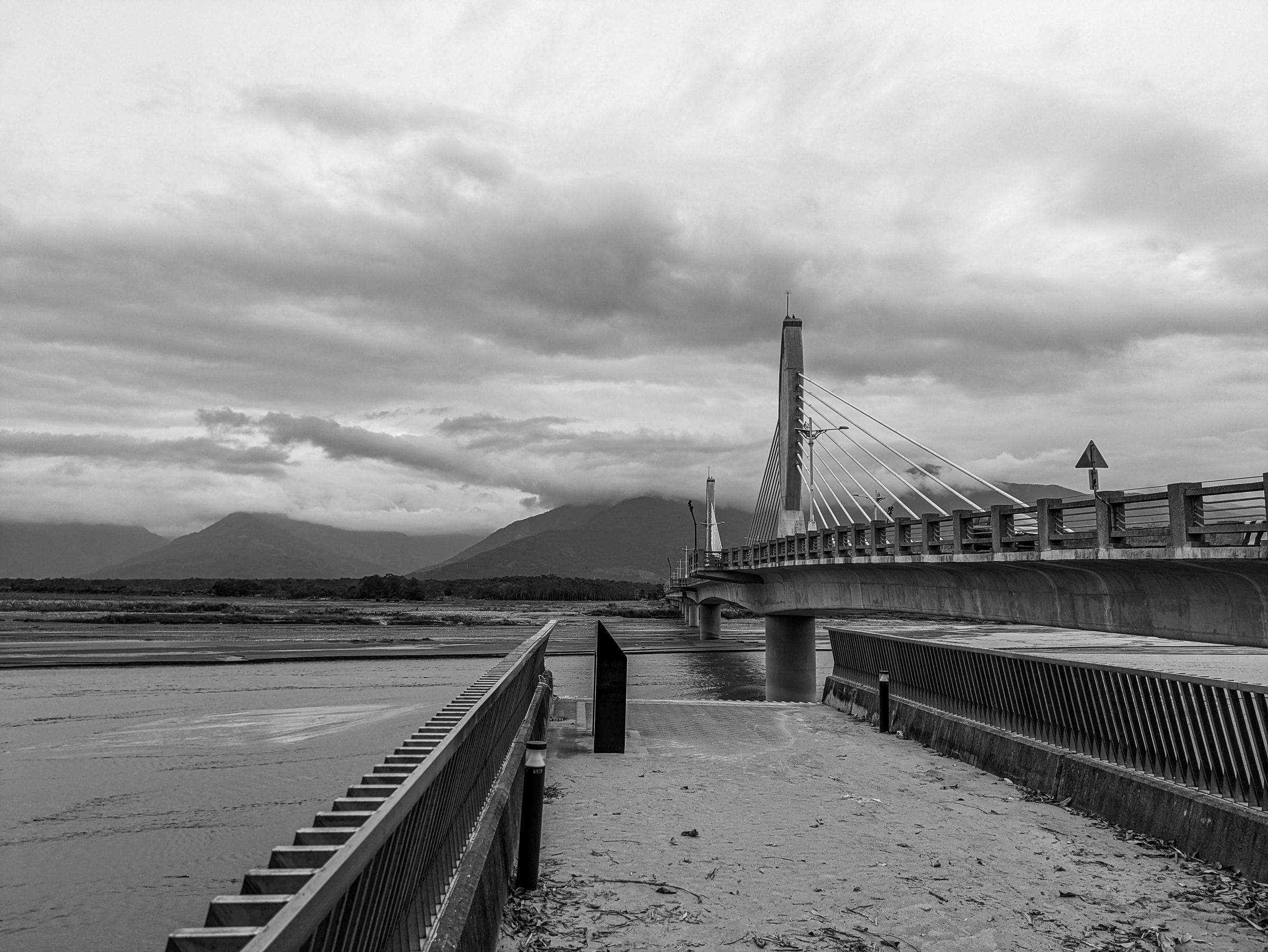 A cable-stayed bridge spans a river with mountains visible under cloudy skies.