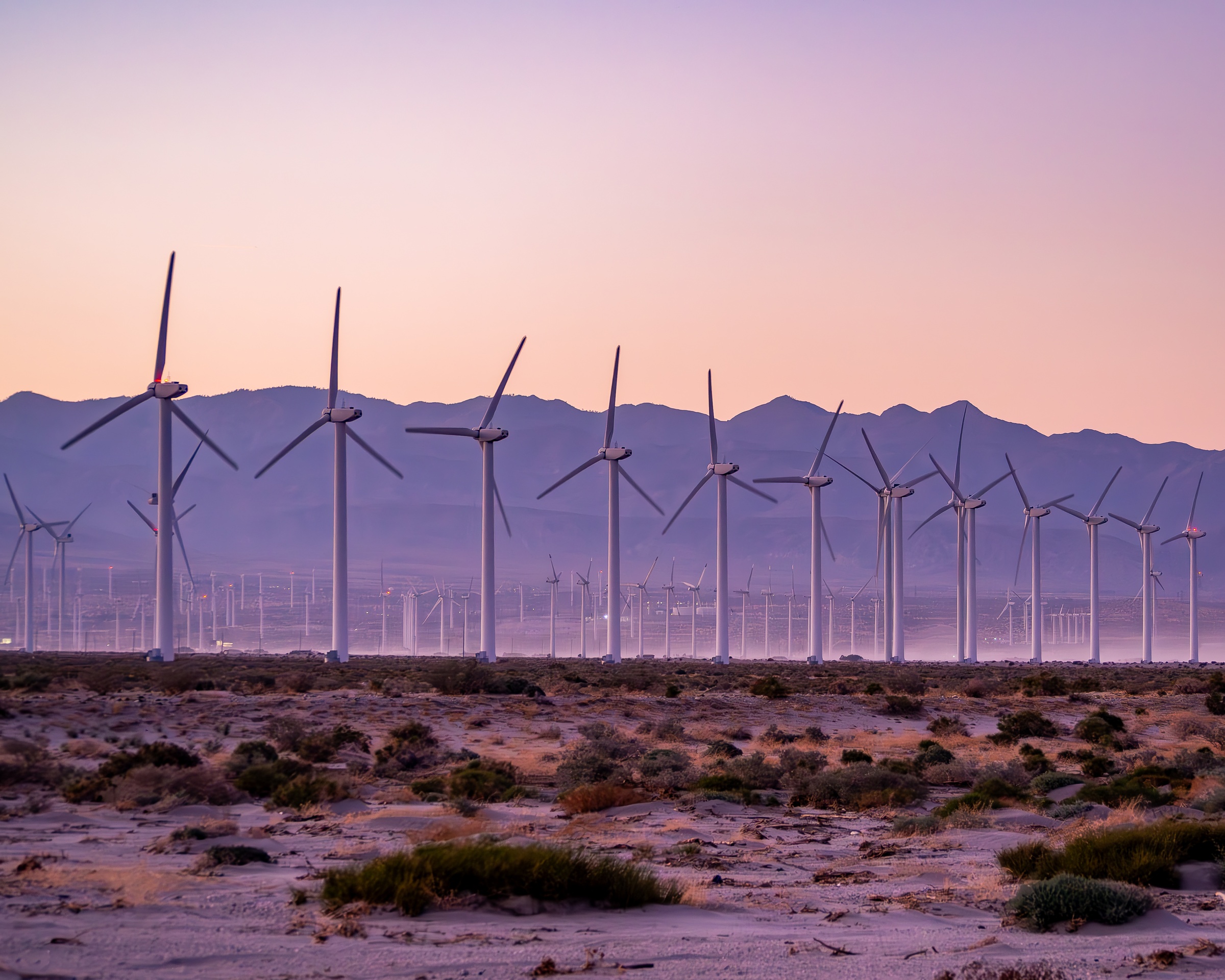 Wind turbines stand in a desert landscape beneath a pastel sky with distant mountains.