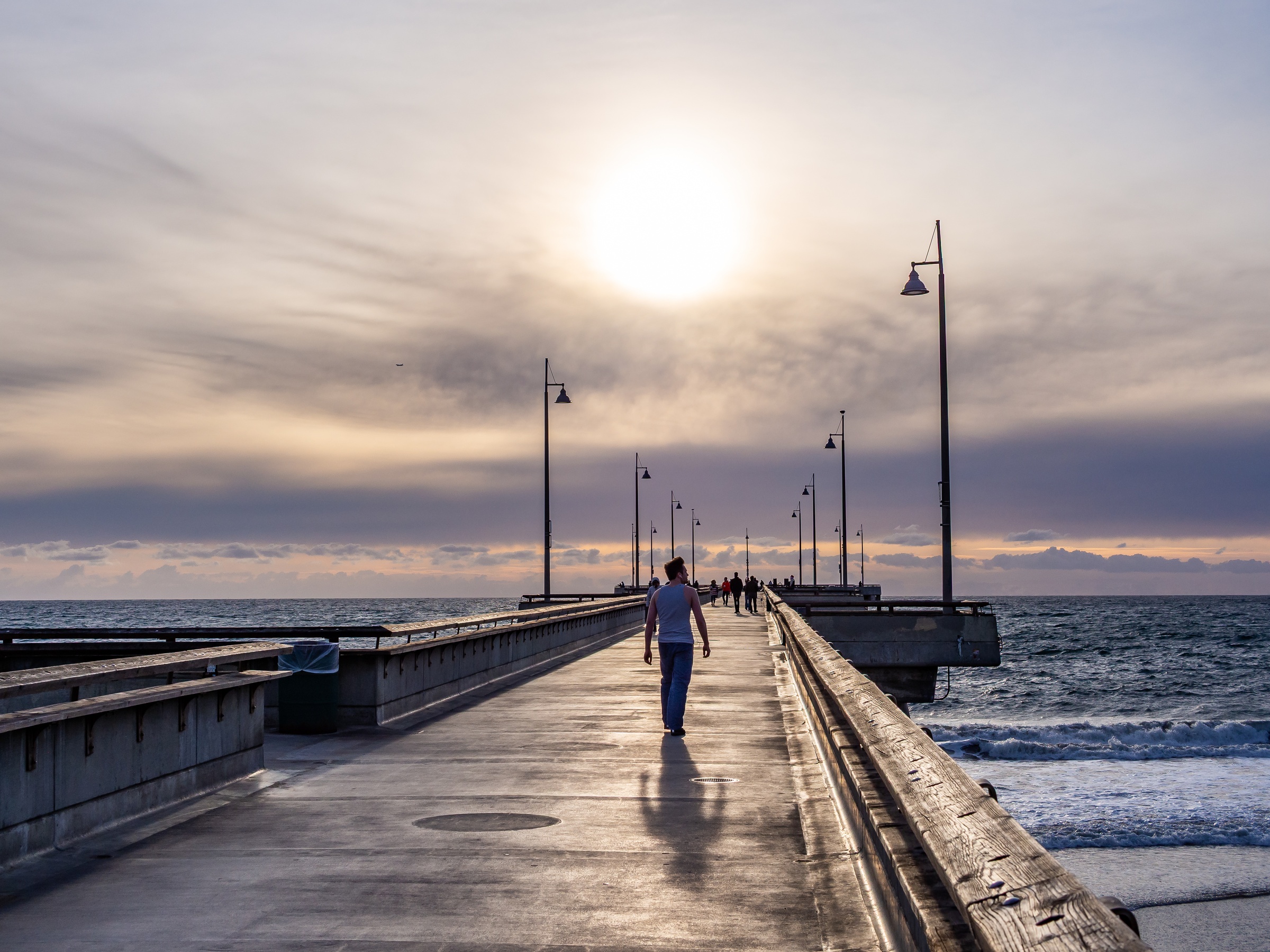 A person walks along a concrete pier toward a bright sun over the ocean.