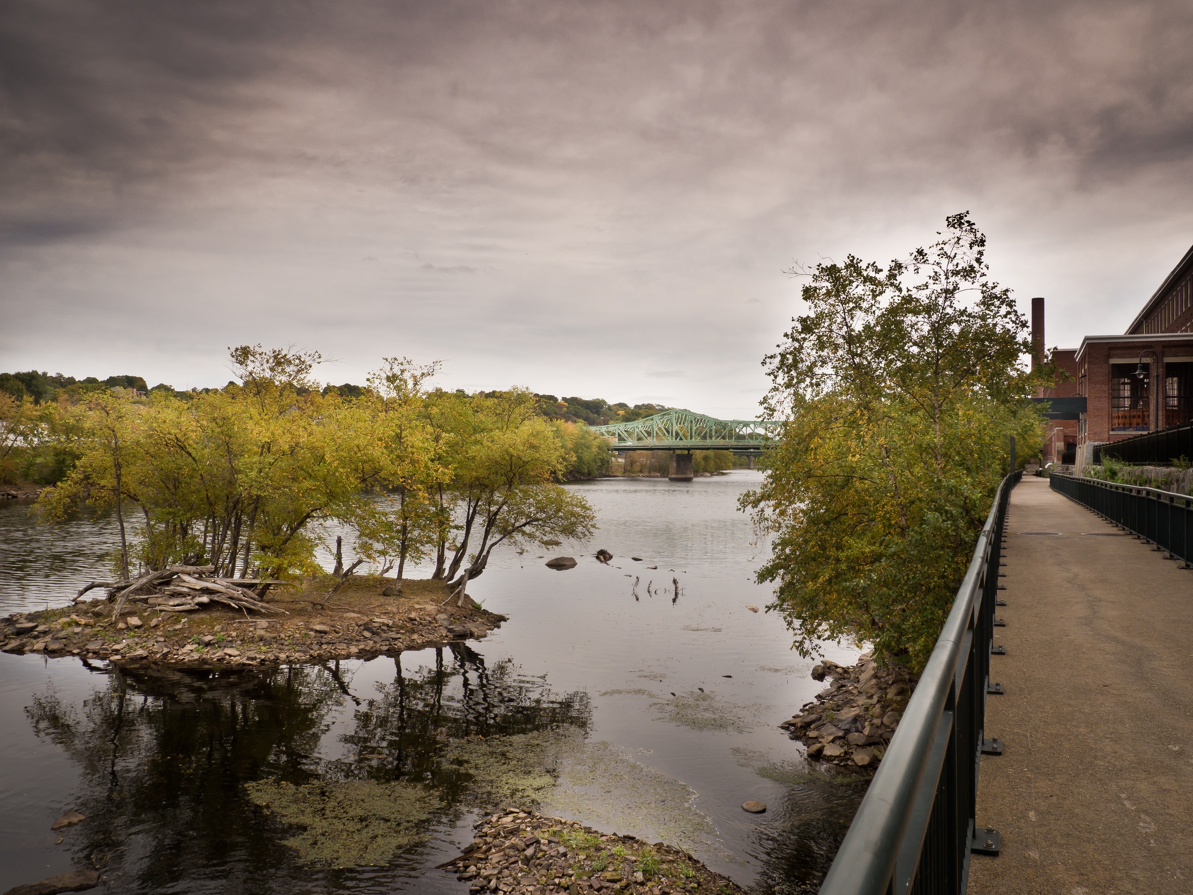 A river flows past trees and a bridge under an overcast sky with a walkway along the bank.