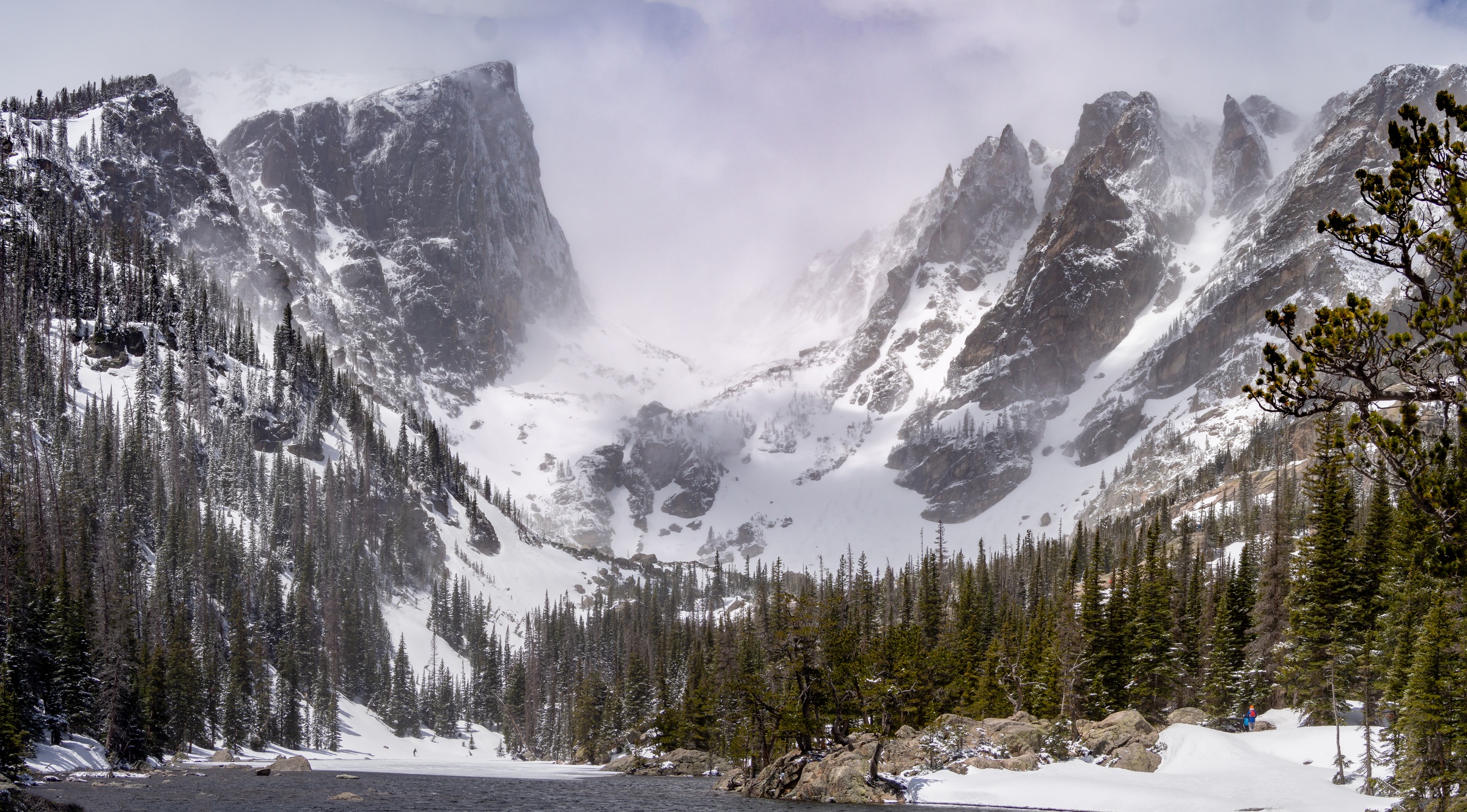 Snow-covered jagged peaks tower over a valley of evergreen trees under a cloudy sky.