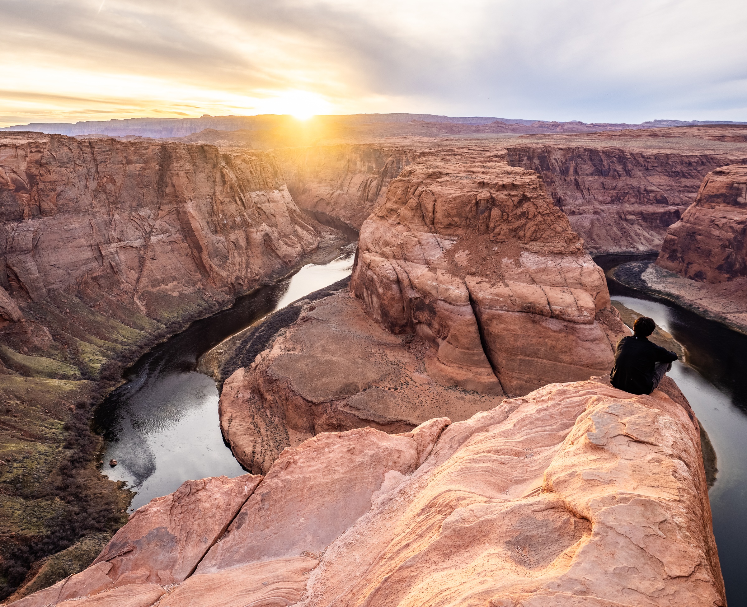 A person sits on a rocky cliff overlooking a winding river in a deep canyon at sunset.