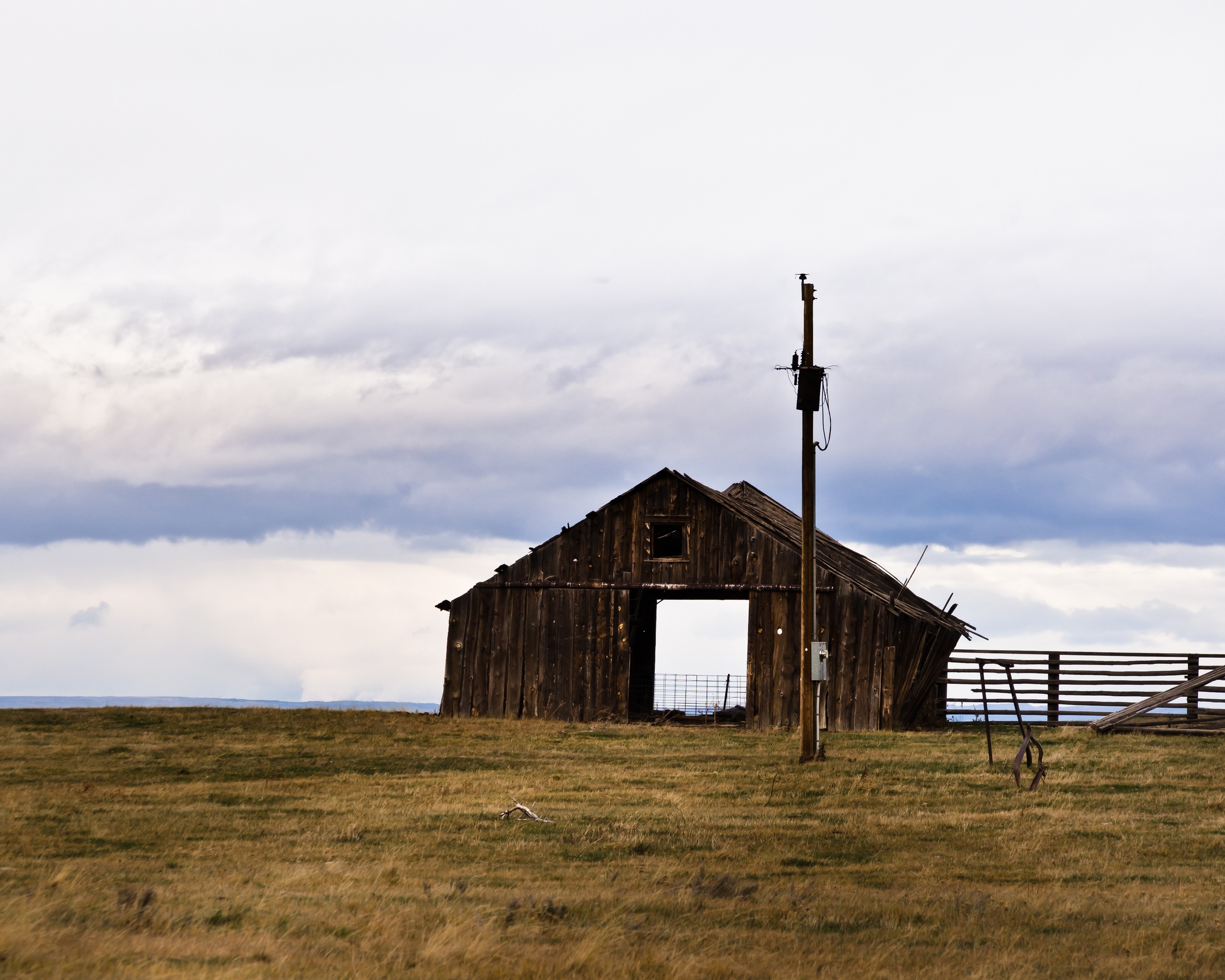 A weathered wooden barn and utility pole stand in a grassy field under a cloudy sky.