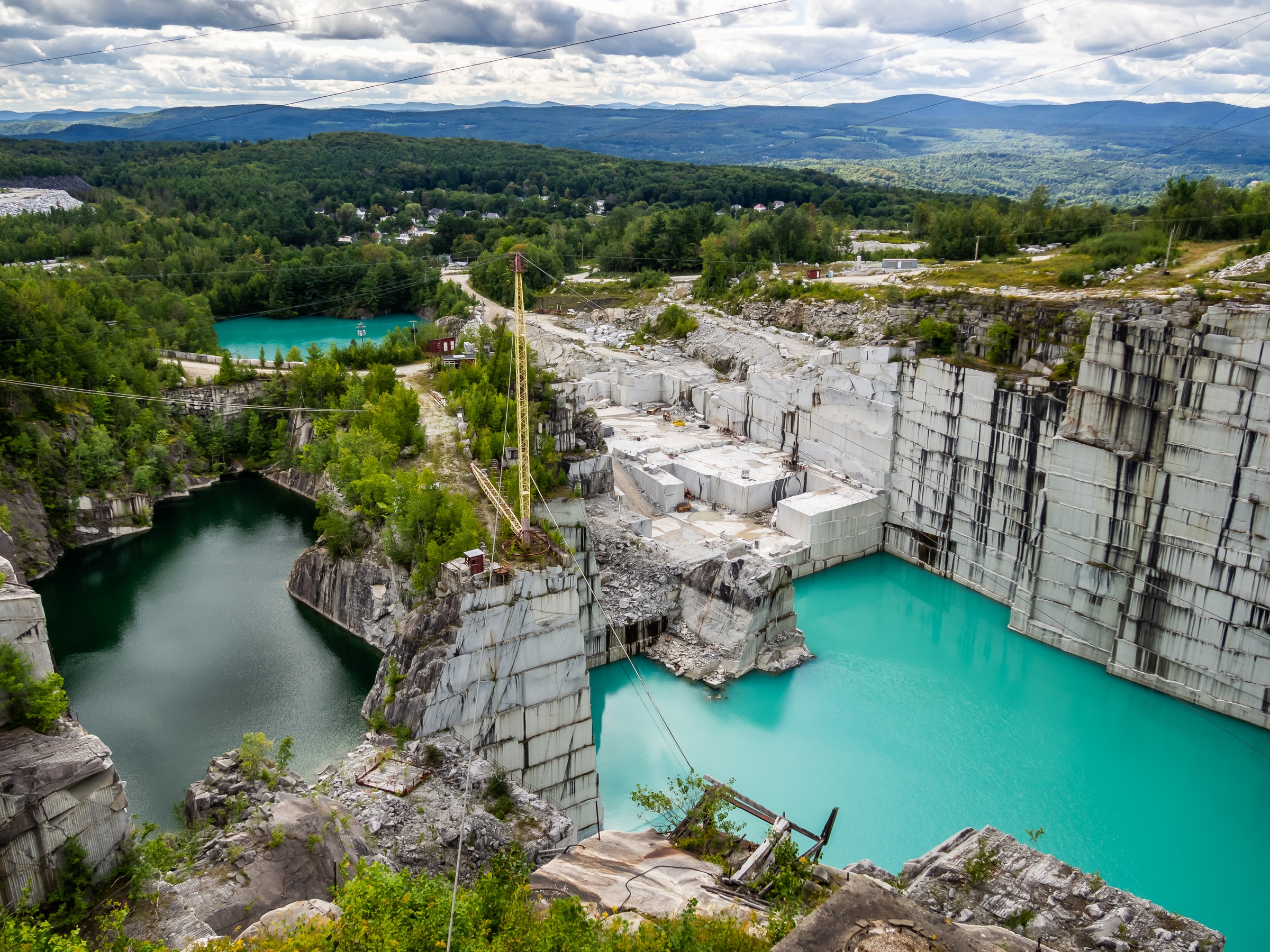 A stone quarry with turquoise water pools and a yellow crane surrounded by green forests.