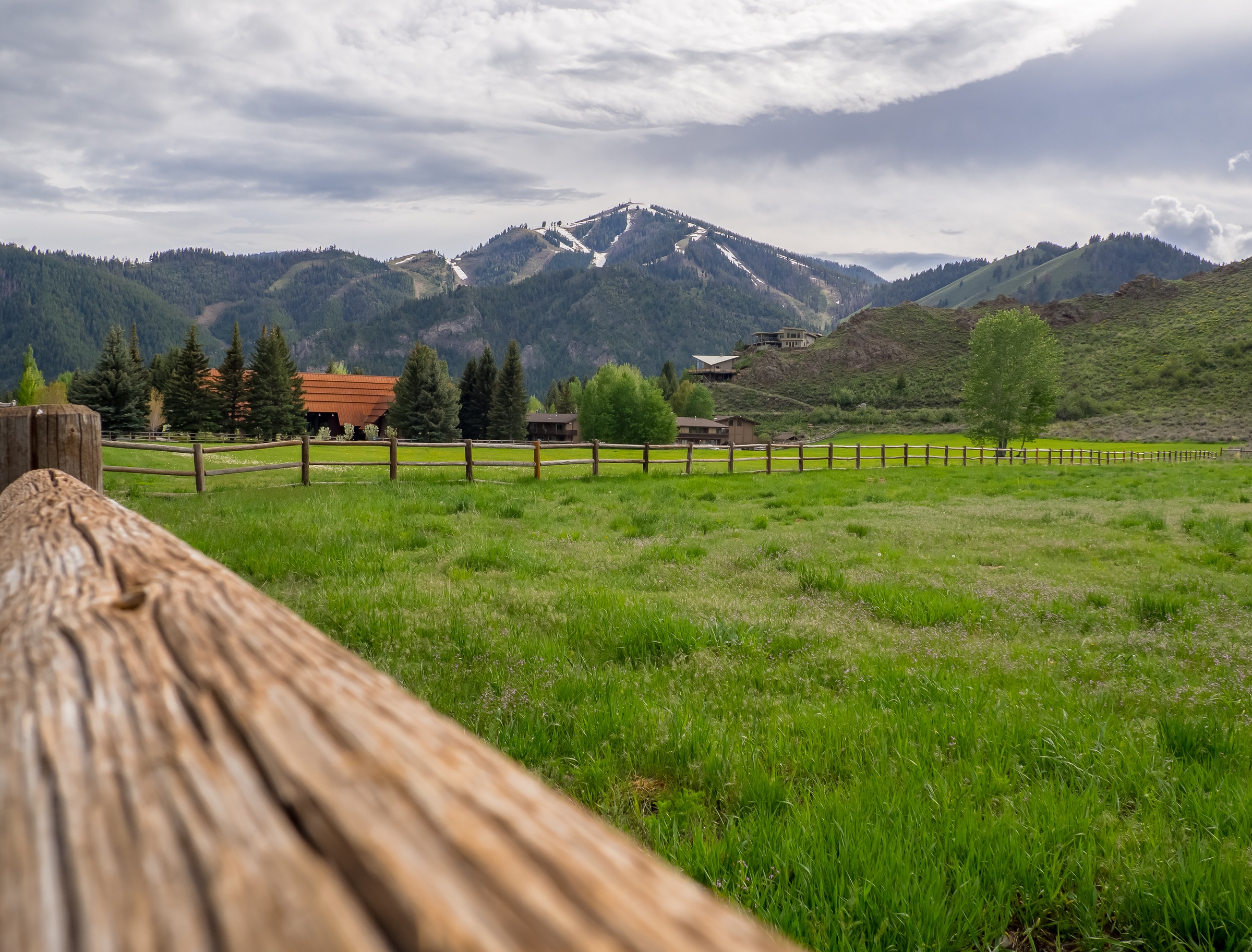 A wooden fence borders a grassy field with buildings and distant mountains under a cloudy sky.