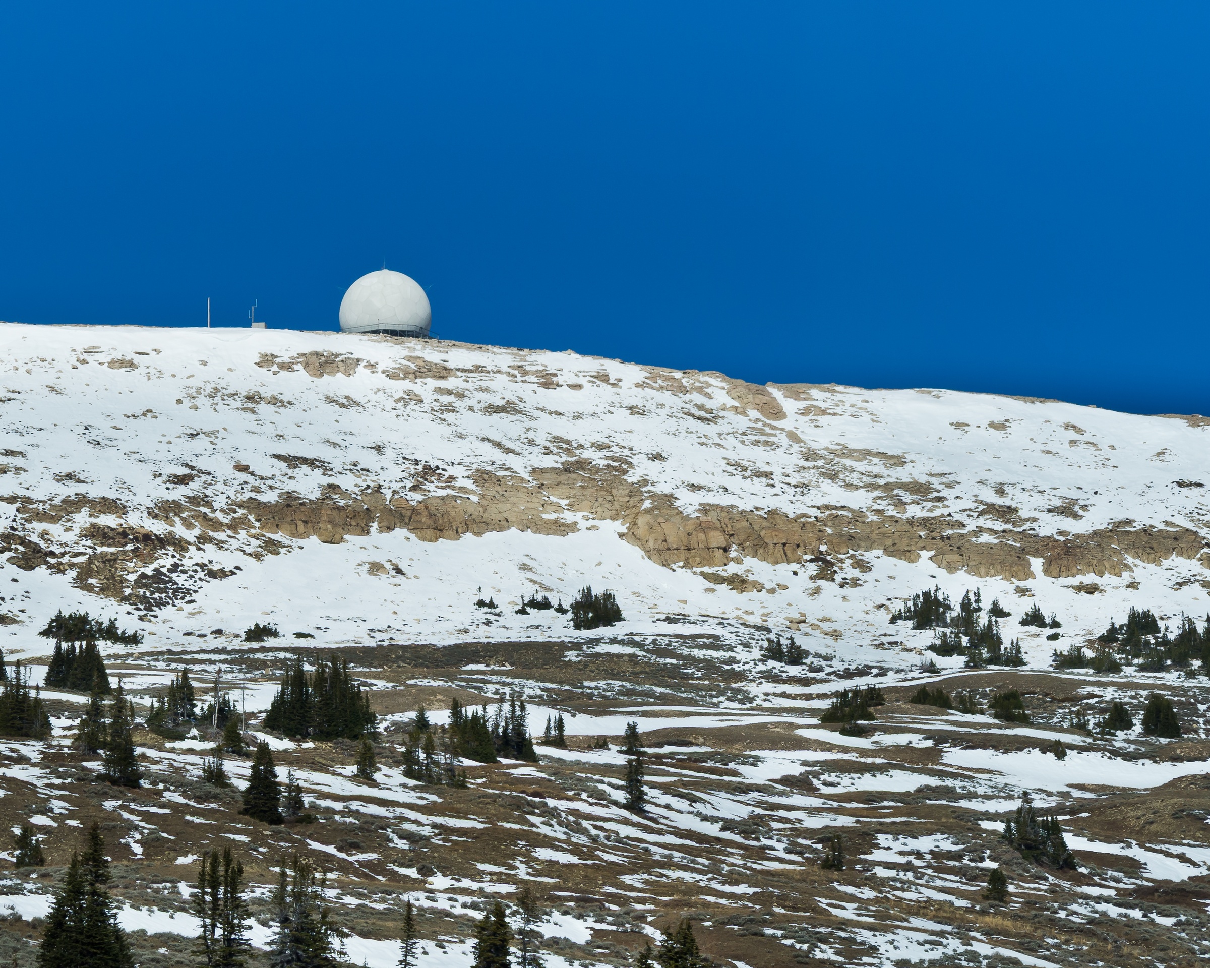 A white spherical structure sits atop a snowy mountain ridge under a clear blue sky.