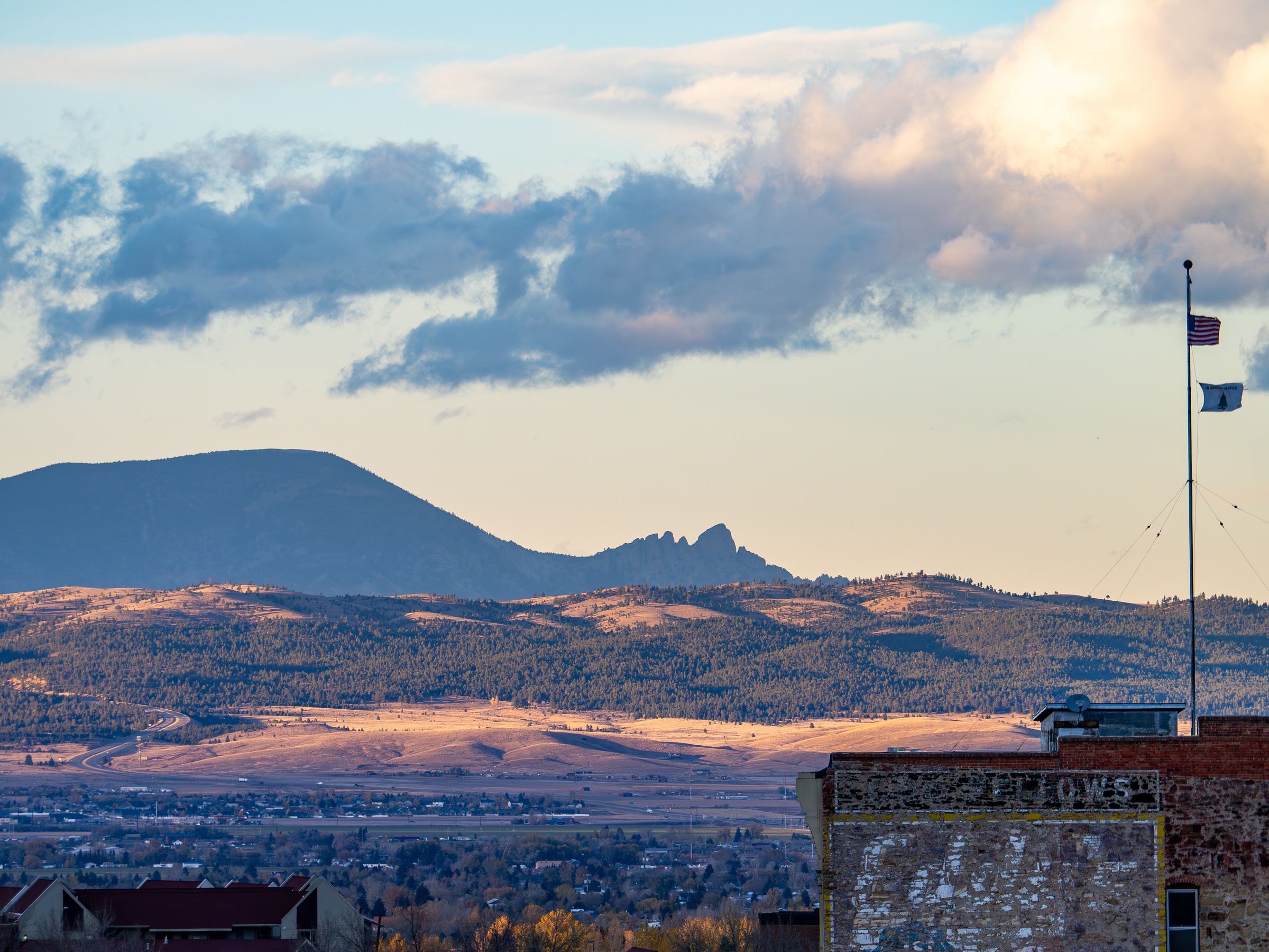 An American flag flies over a town set against rolling hills and distant mountains under clouds.