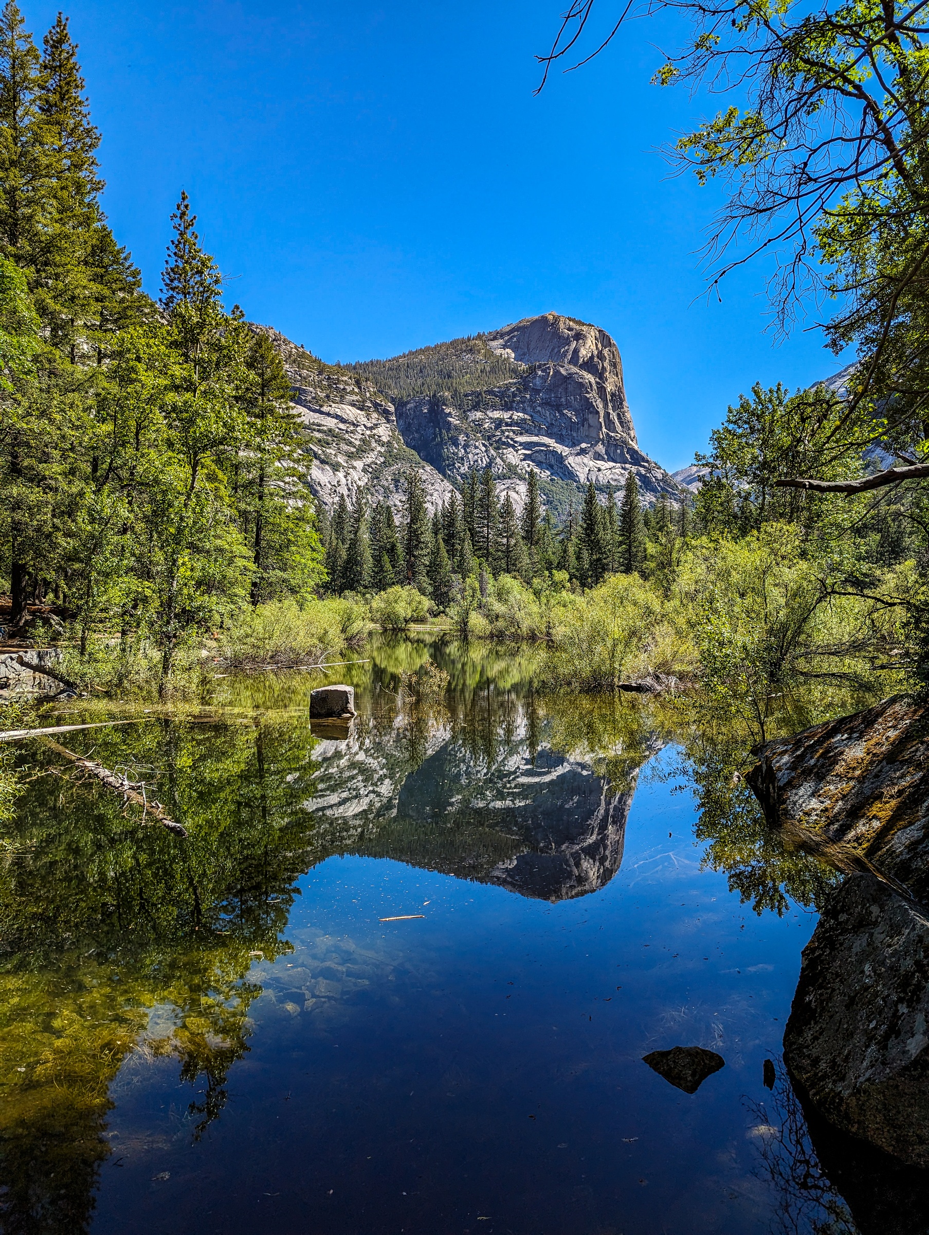 A granite cliff and evergreen trees reflect in a calm lake under a clear blue sky.