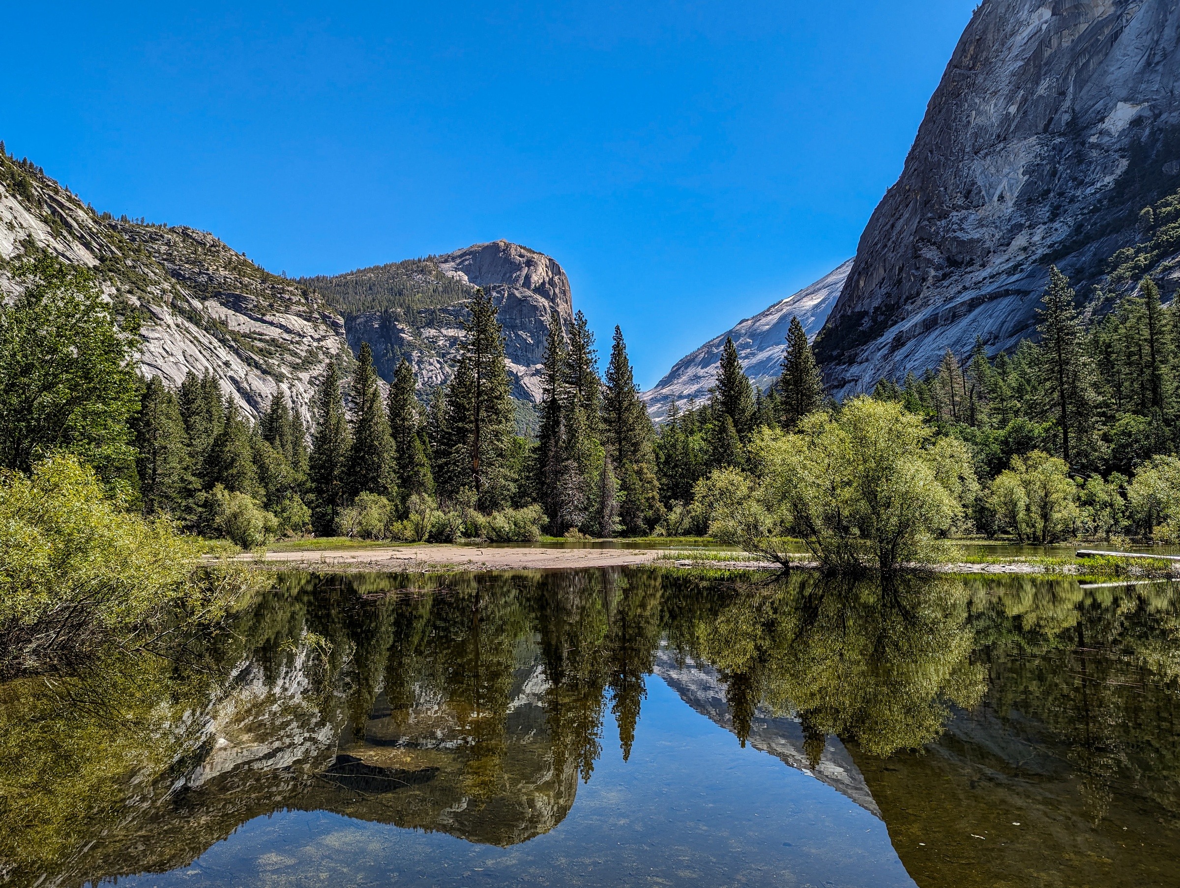 Still water reflects towering granite cliffs and evergreen forests under a clear blue sky.