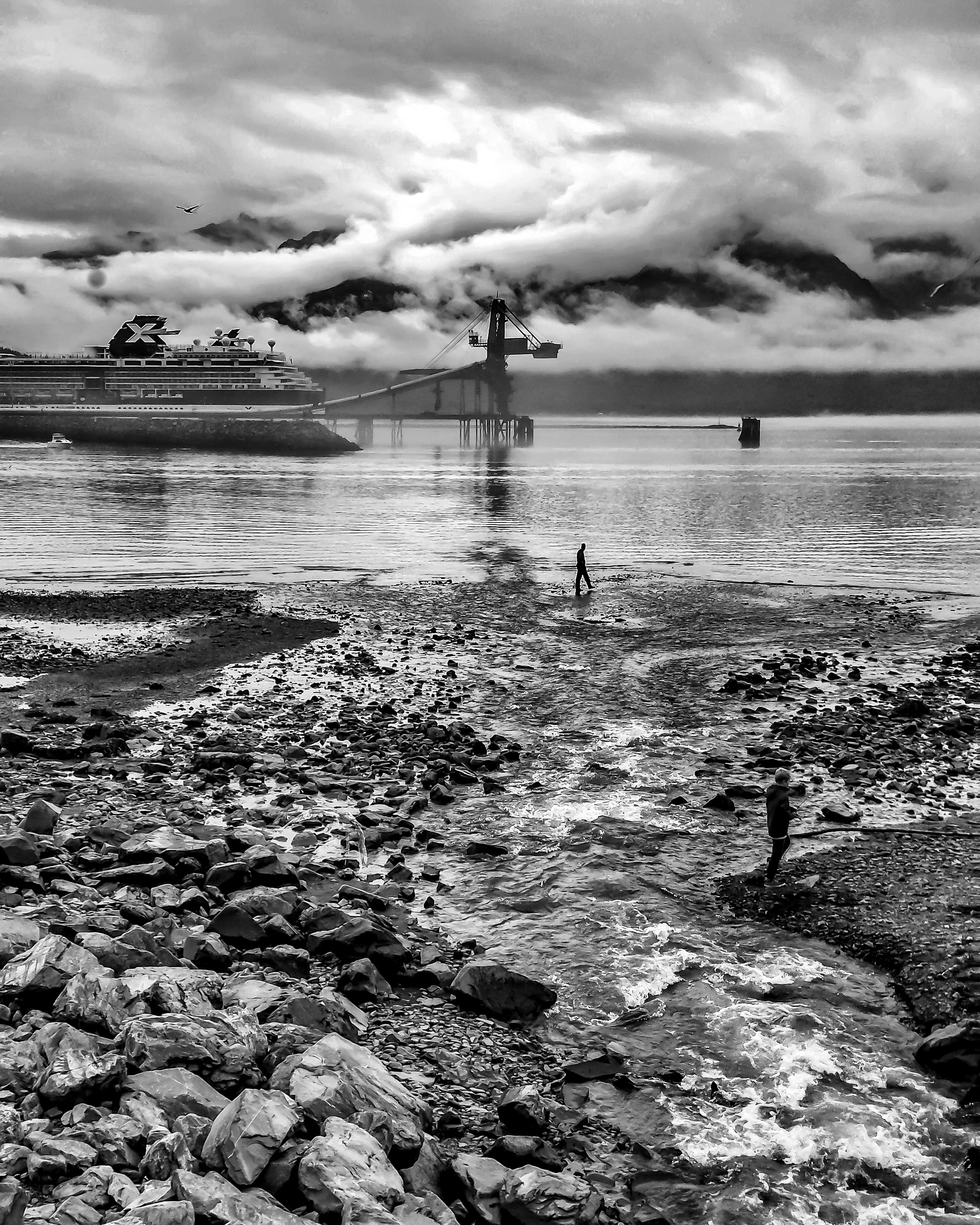 A person stands in shallow water on a rocky shore near a docked cruise ship and mountains.