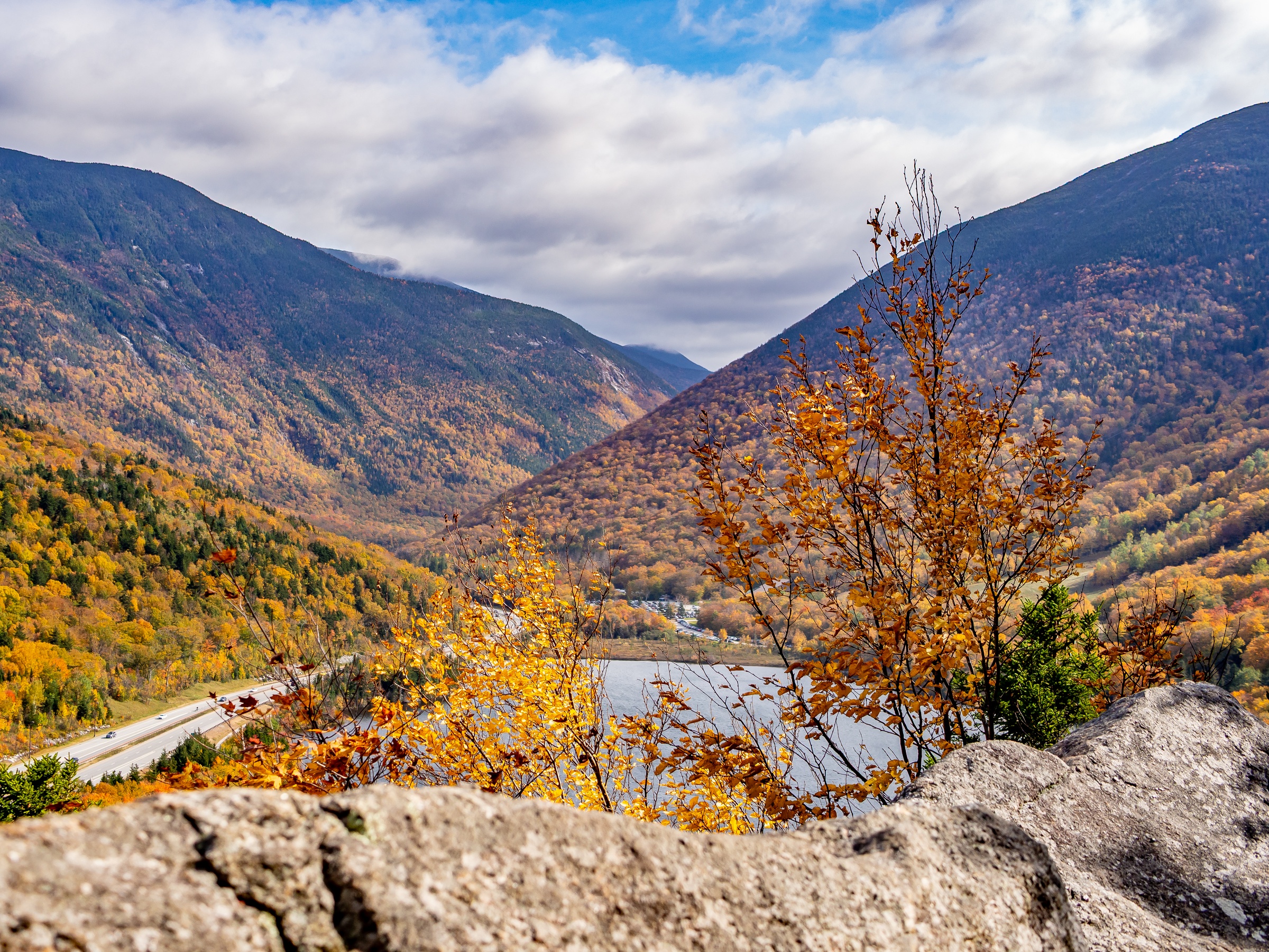 Autumn-colored mountains surround a valley with a river and road viewed from a rocky overlook.