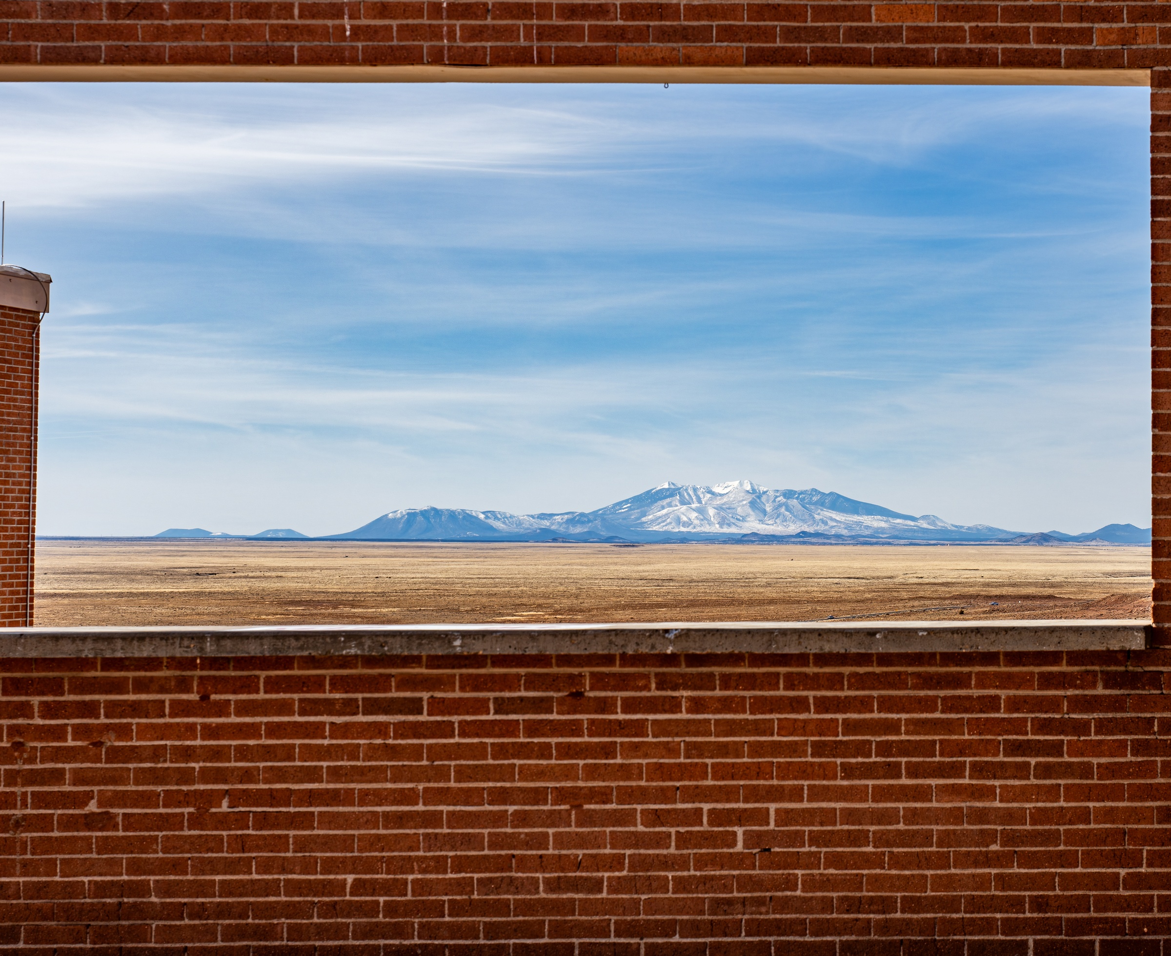 Snowy mountains and flat plains seen through a rectangular opening in a brick wall.