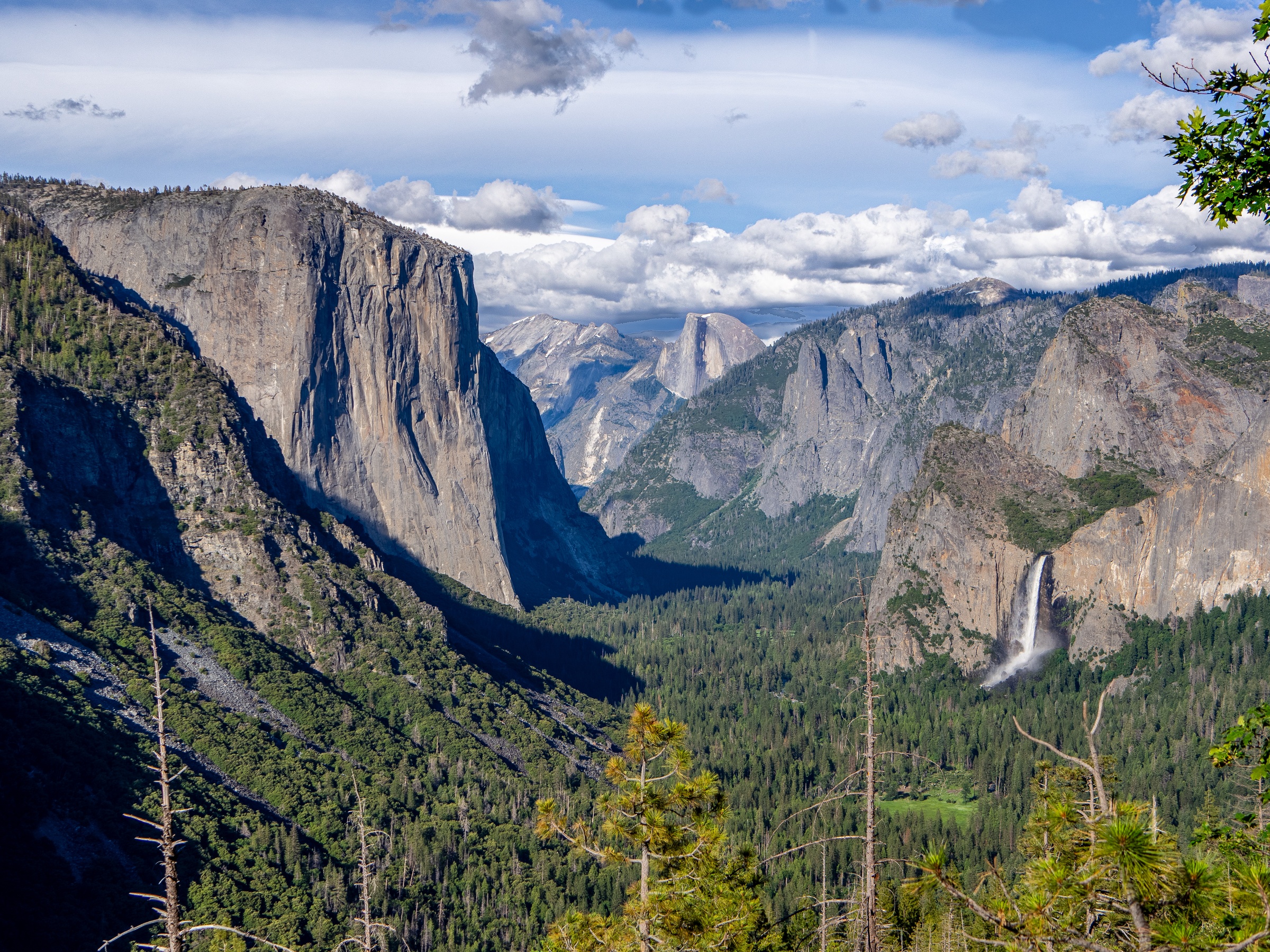 Towering granite cliffs and a waterfall surround a forested valley under a cloudy sky.