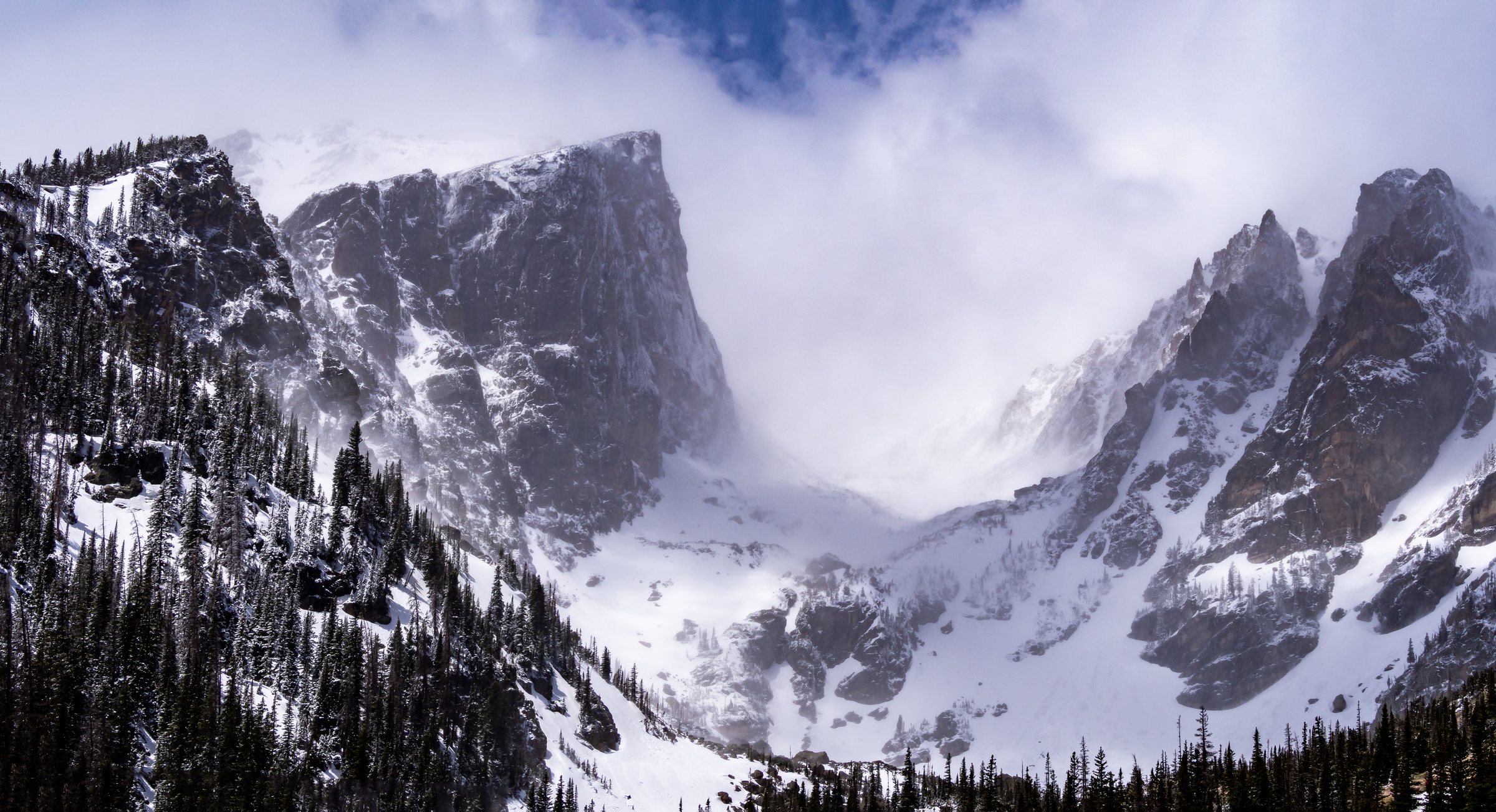 Snowy mountain cliffs and evergreen trees surround a valley filled with white clouds.