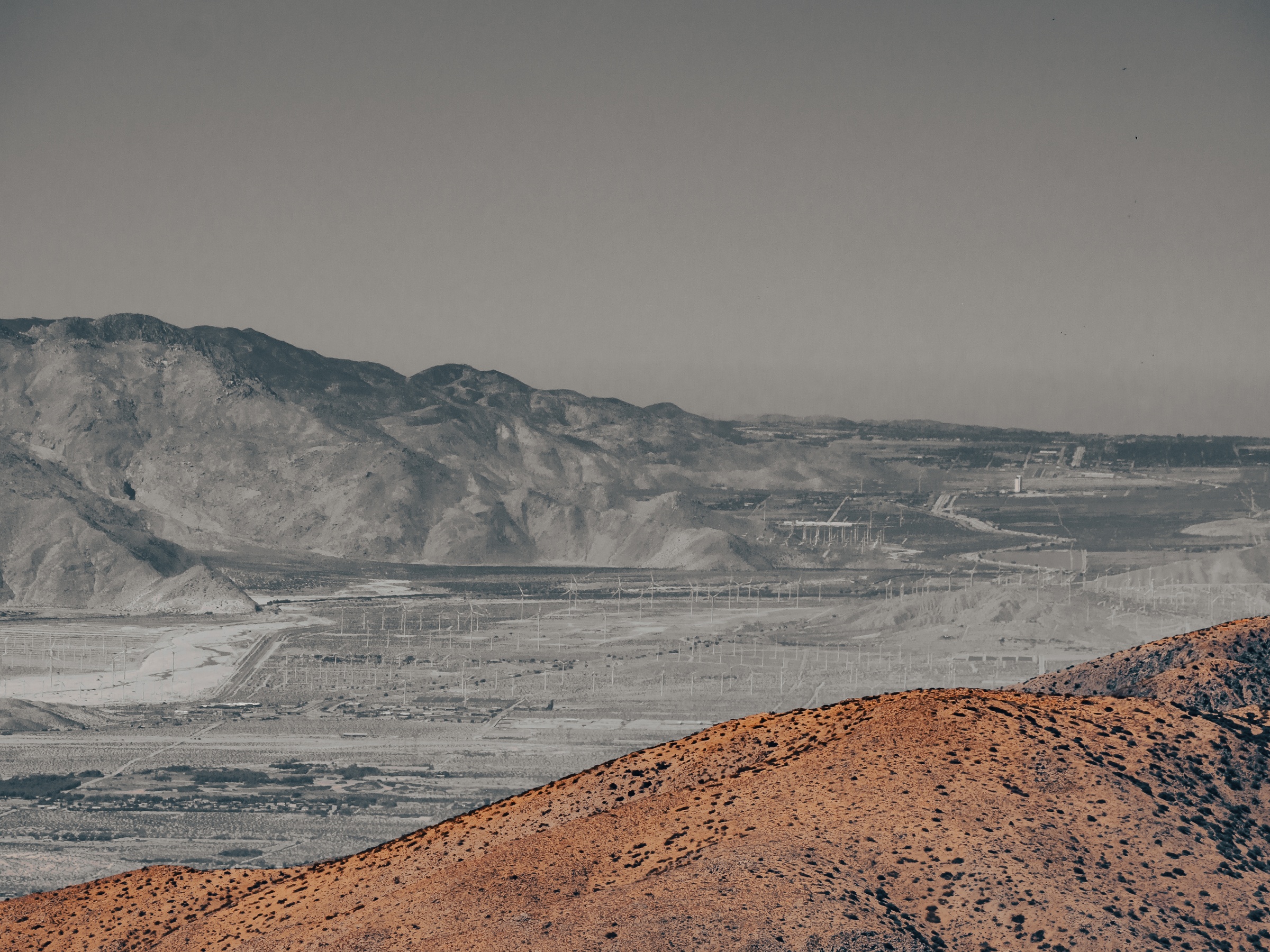 Brown hills overlook a wide, pale valley and distant mountains under a grey sky.