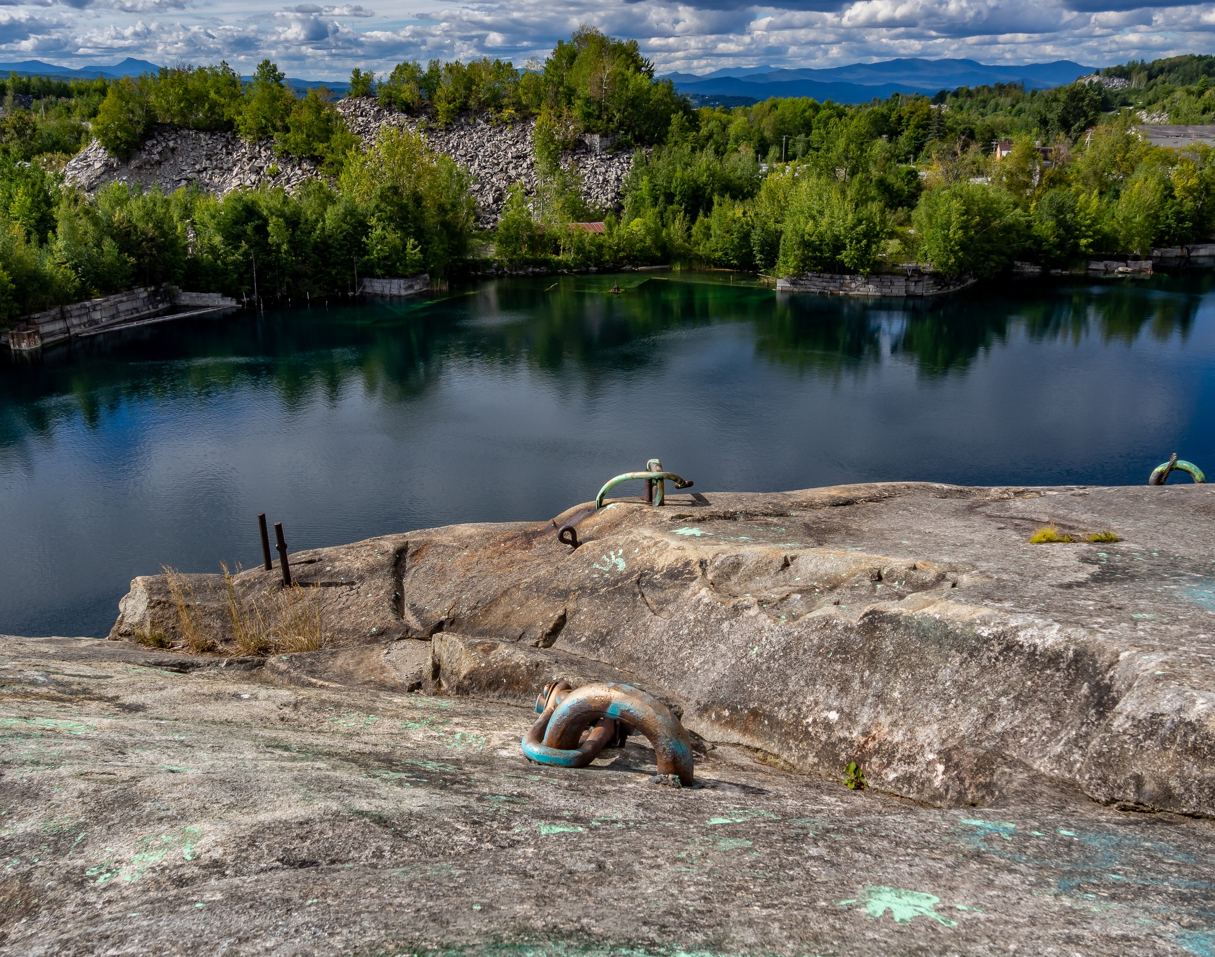 Metal pipes rest on rocky terrain overlooking a blue lake surrounded by green trees and mountains.