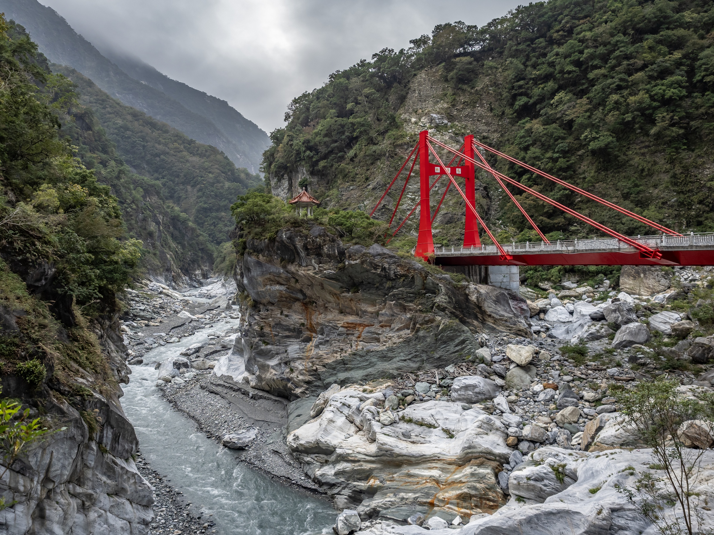 A red bridge spans a rocky riverbed between steep, forested mountains.