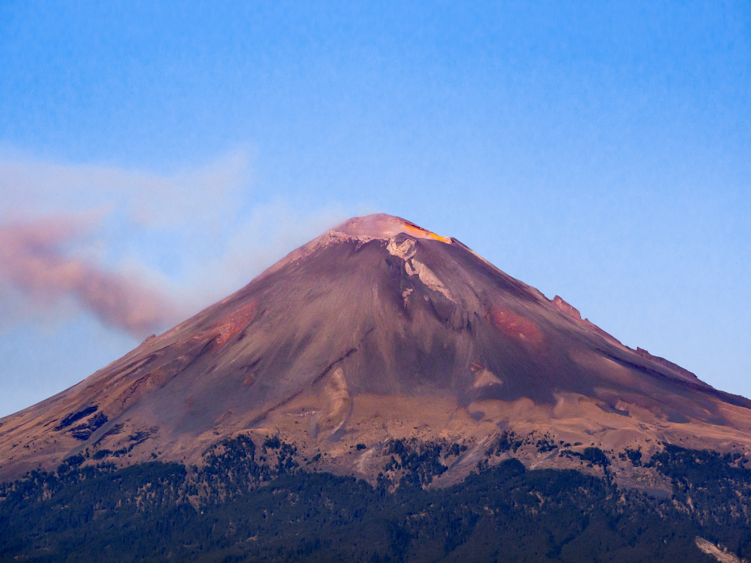A smoking volcano rises above a forest under a clear blue sky.