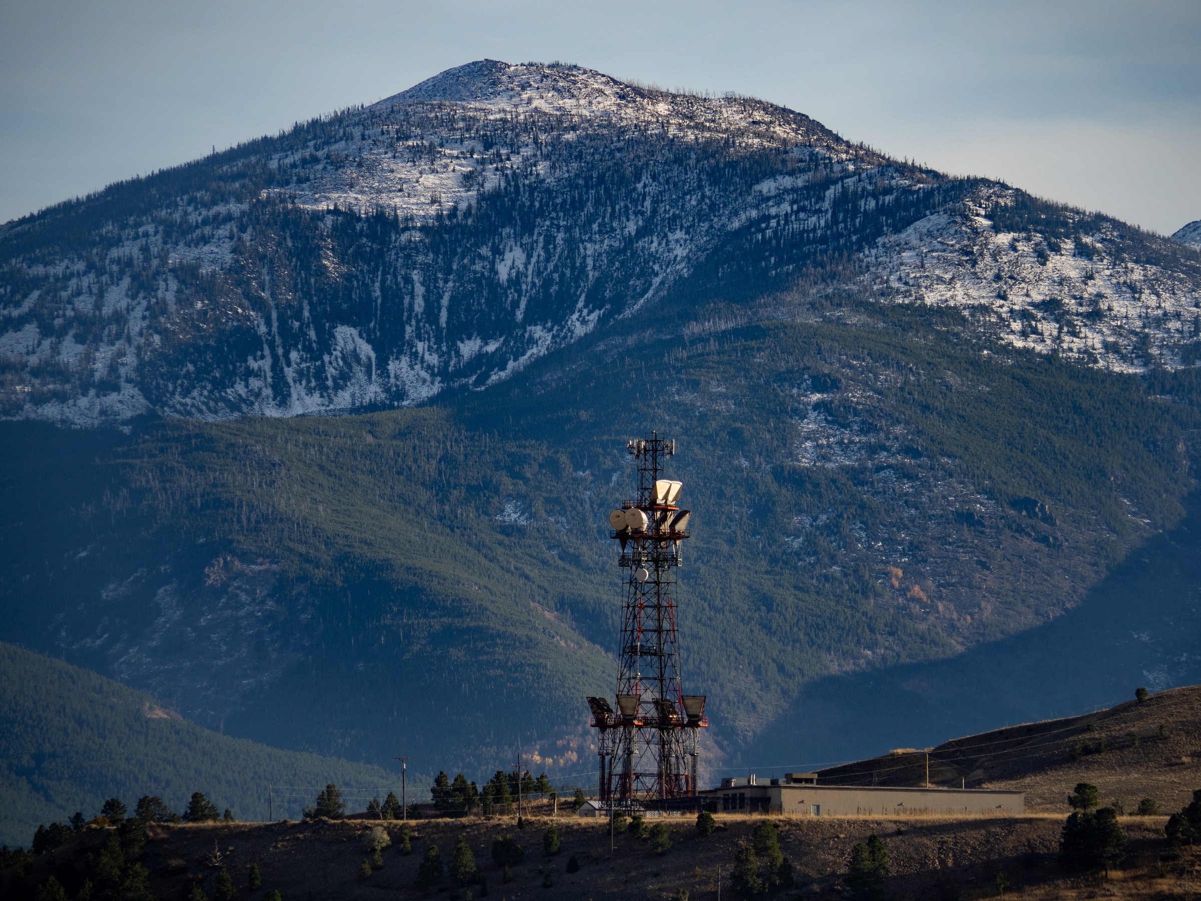 A communication tower stands before a large, snow-dusted mountain.