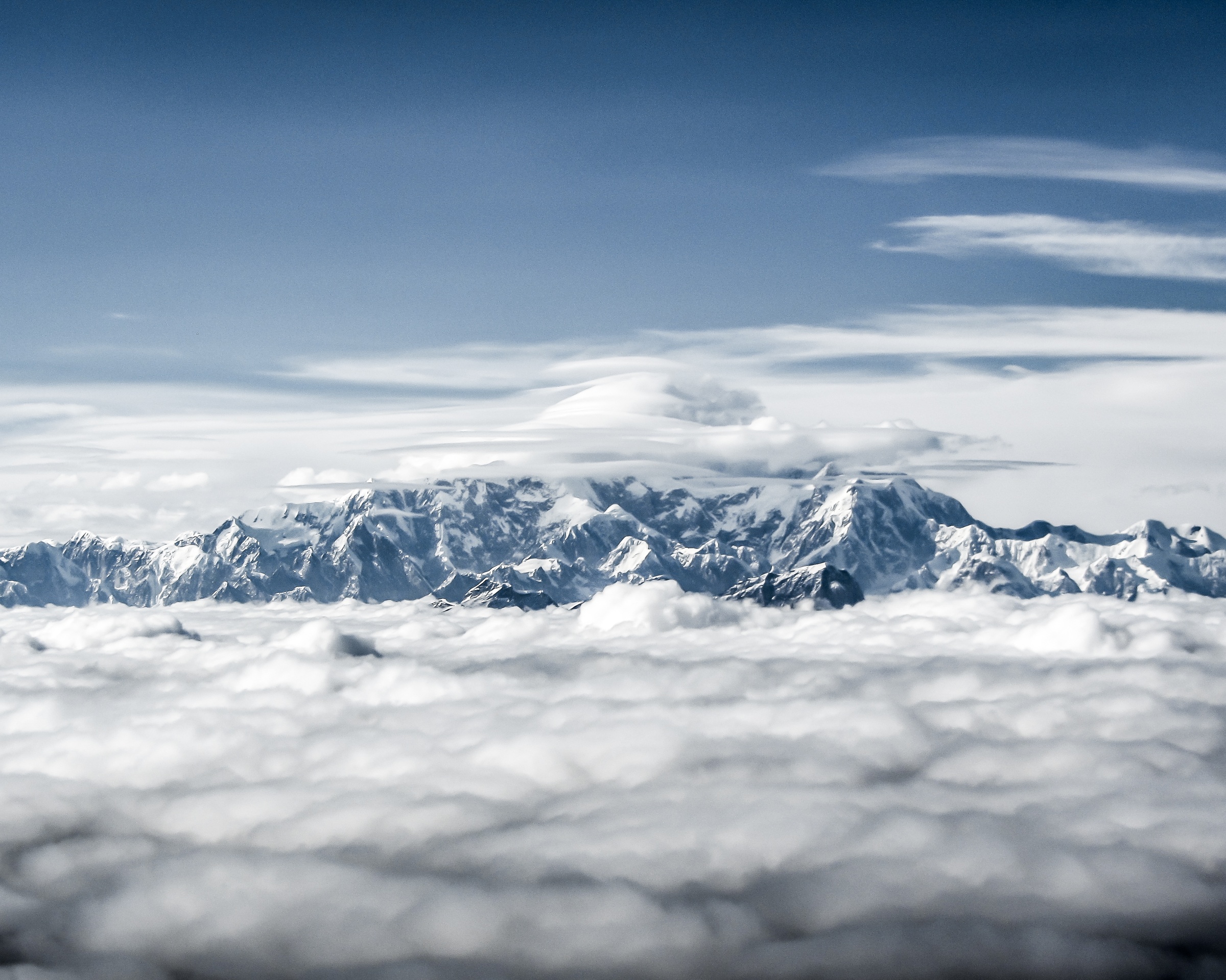 Snow-capped mountains rise above a thick layer of white clouds under a clear blue sky.