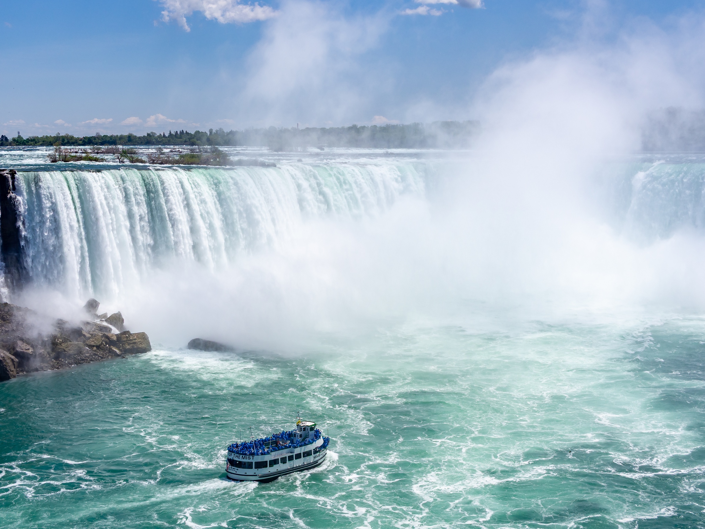 A tour boat floats on turquoise water at the base of a massive, misty waterfall.