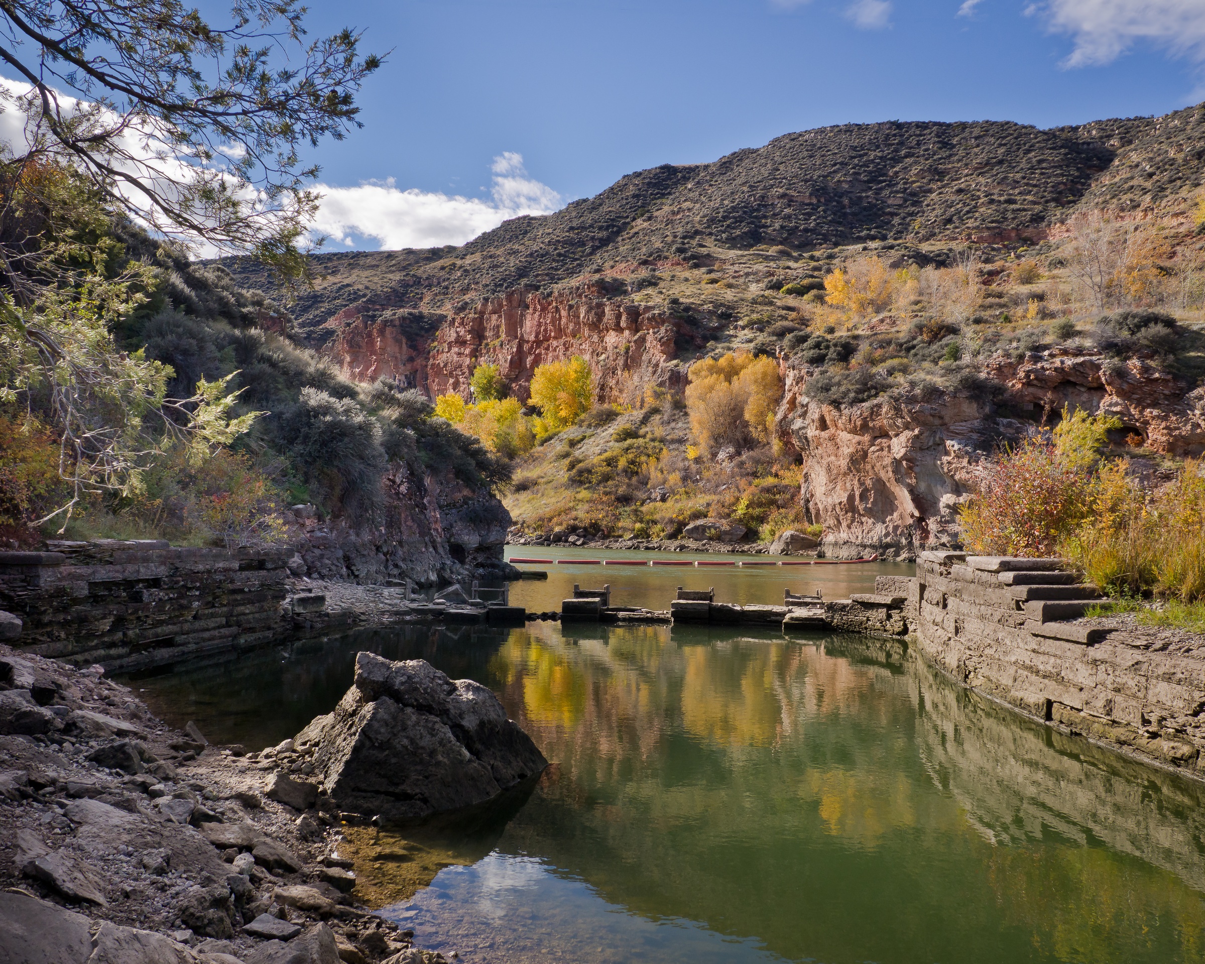 A reflective river with stone structures is surrounded by autumn foliage and rugged brown mountains.