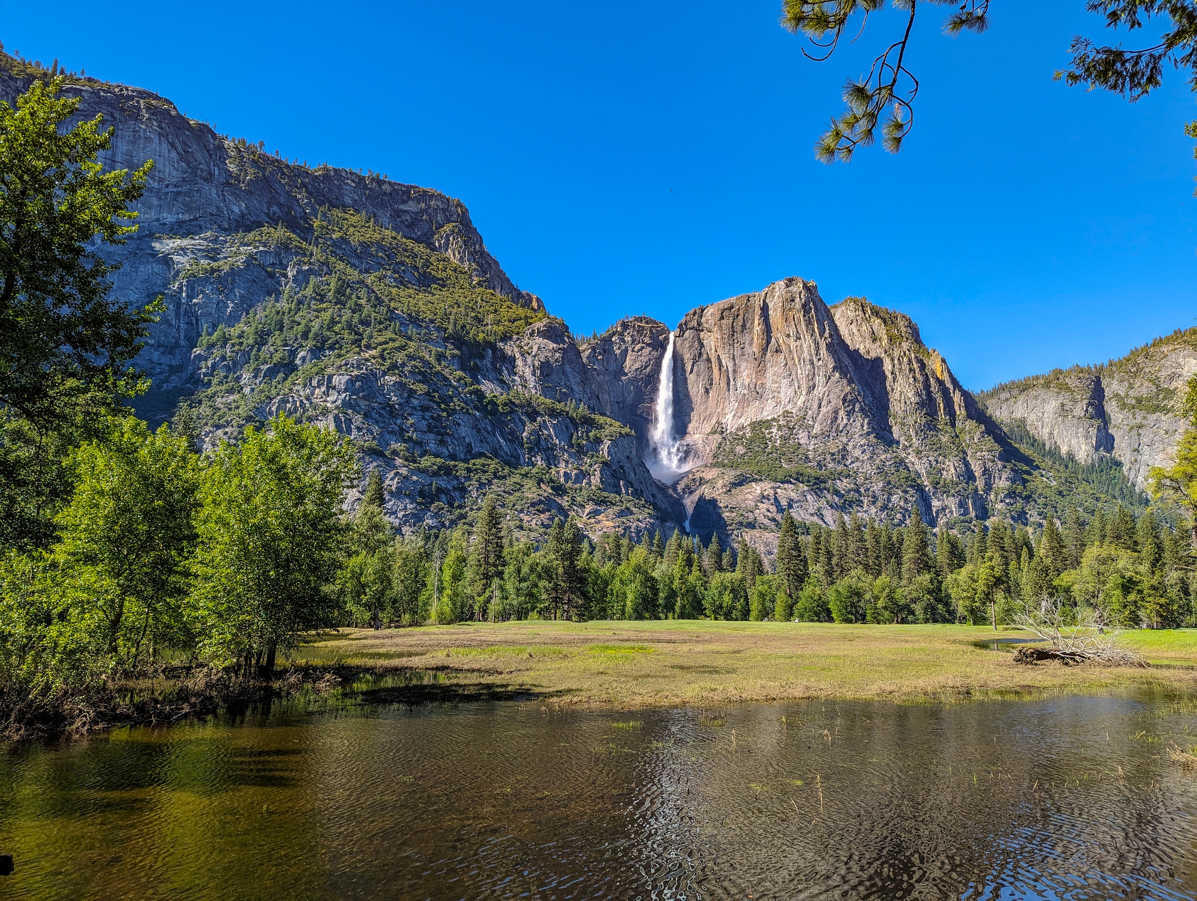 A tall waterfall cascades down granite cliffs above a forested valley and a still pond.