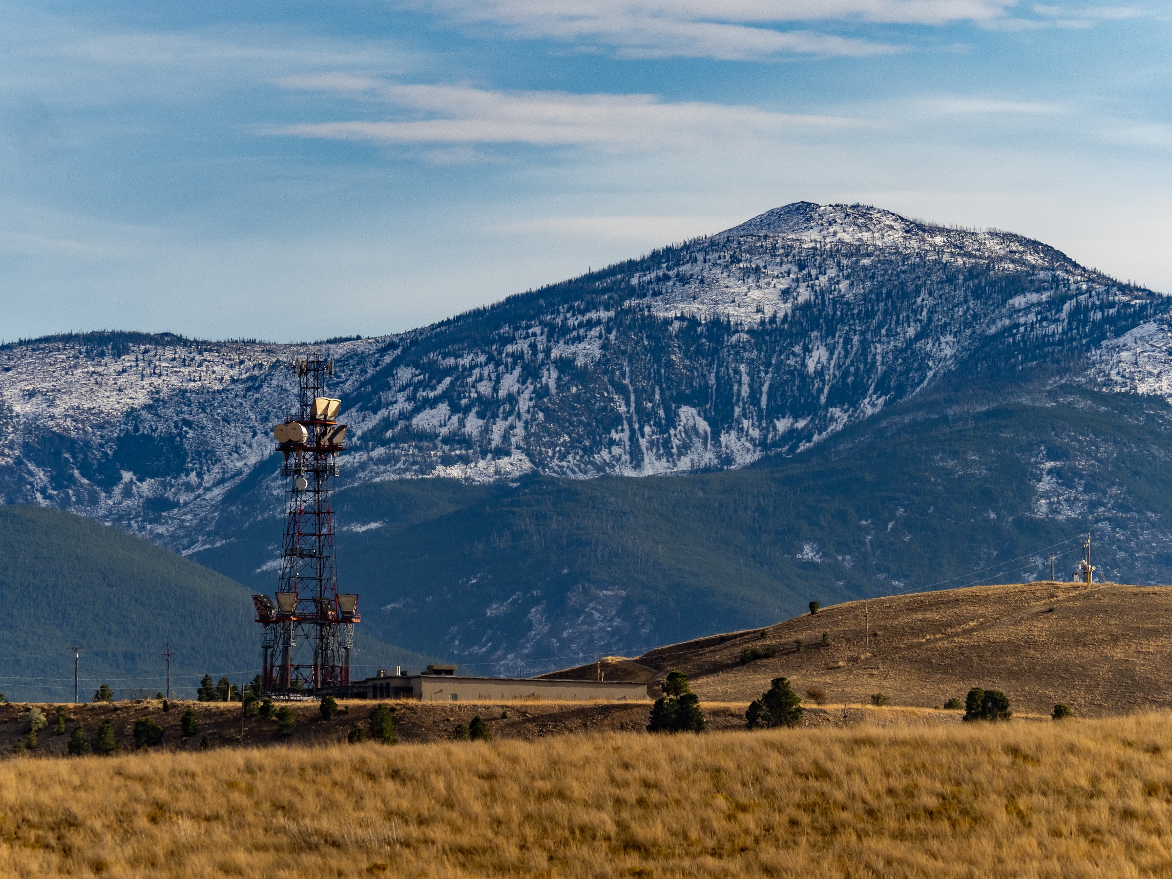 A communication tower stands in a grassy field before snow-covered mountains under a blue sky.