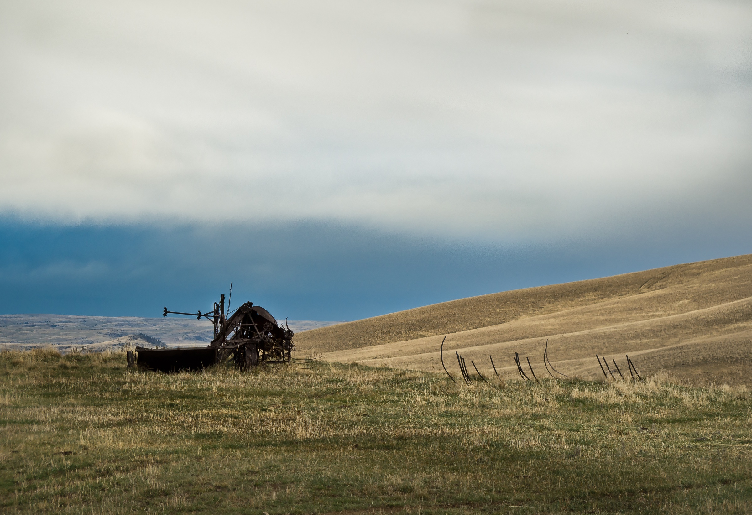 Rusty farm machinery rests in a grassy field with rolling hills under a cloudy sky.