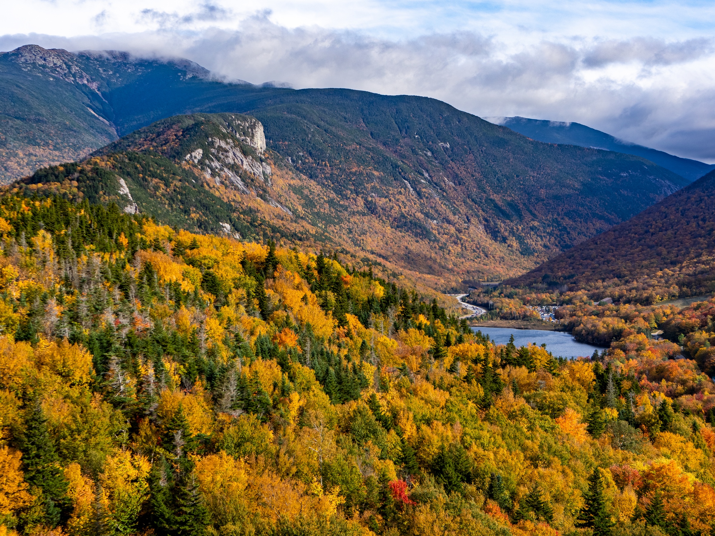 Autumn-colored trees cover mountains surrounding a small lake under a cloudy sky.