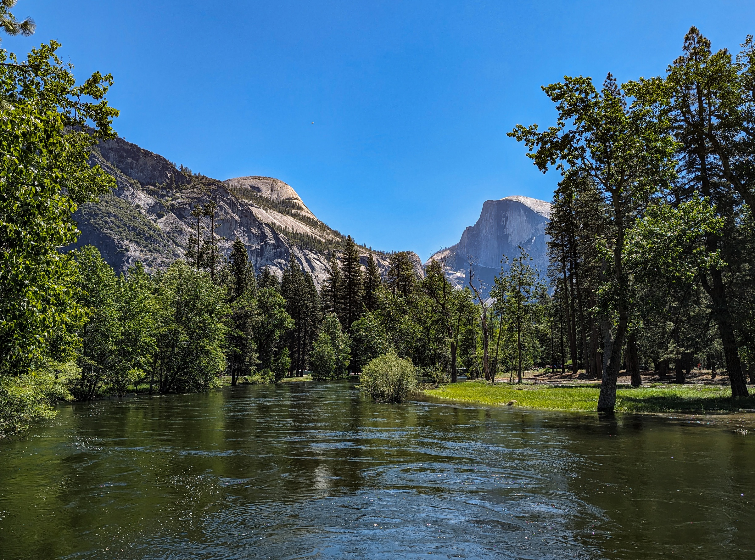 A river flows through a green valley beneath towering granite mountains and a clear blue sky.