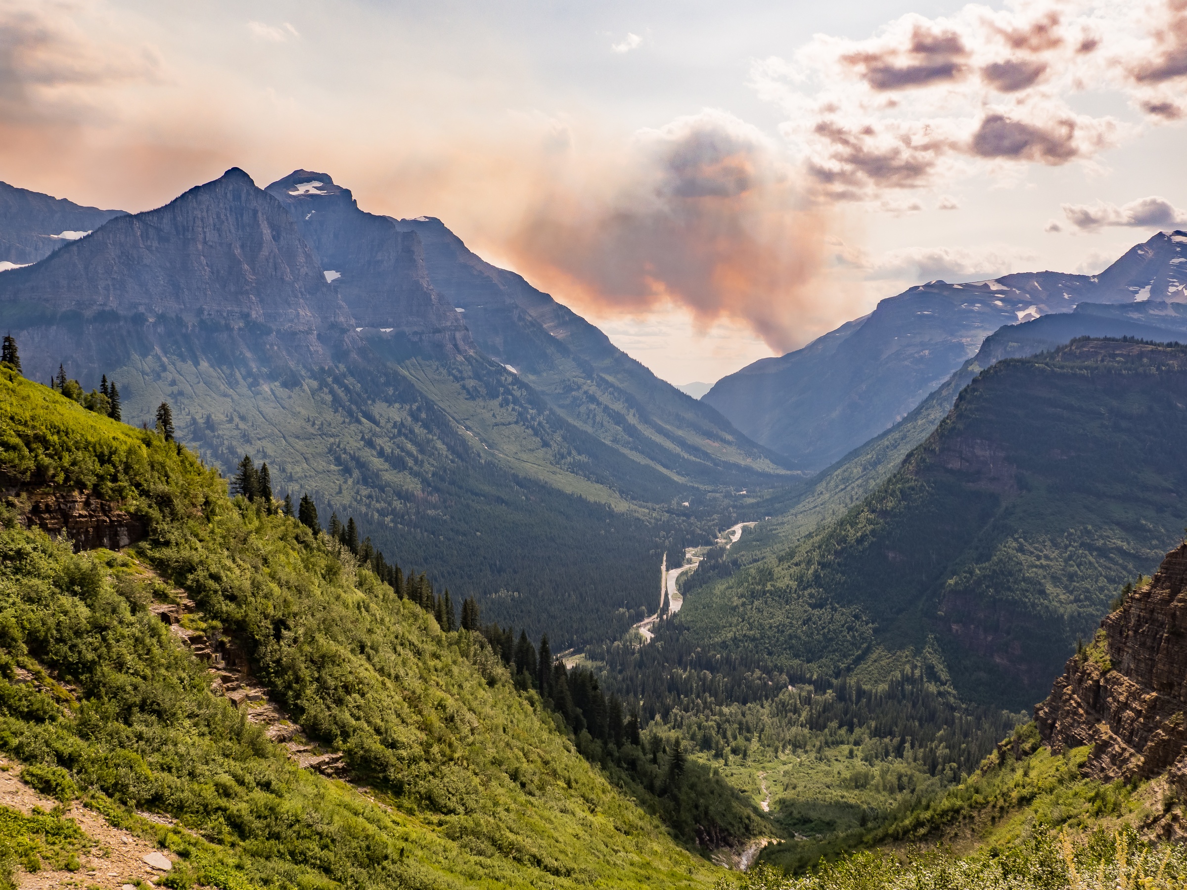 A winding road runs through a lush mountain valley beneath a hazy, smoke-filled sky.