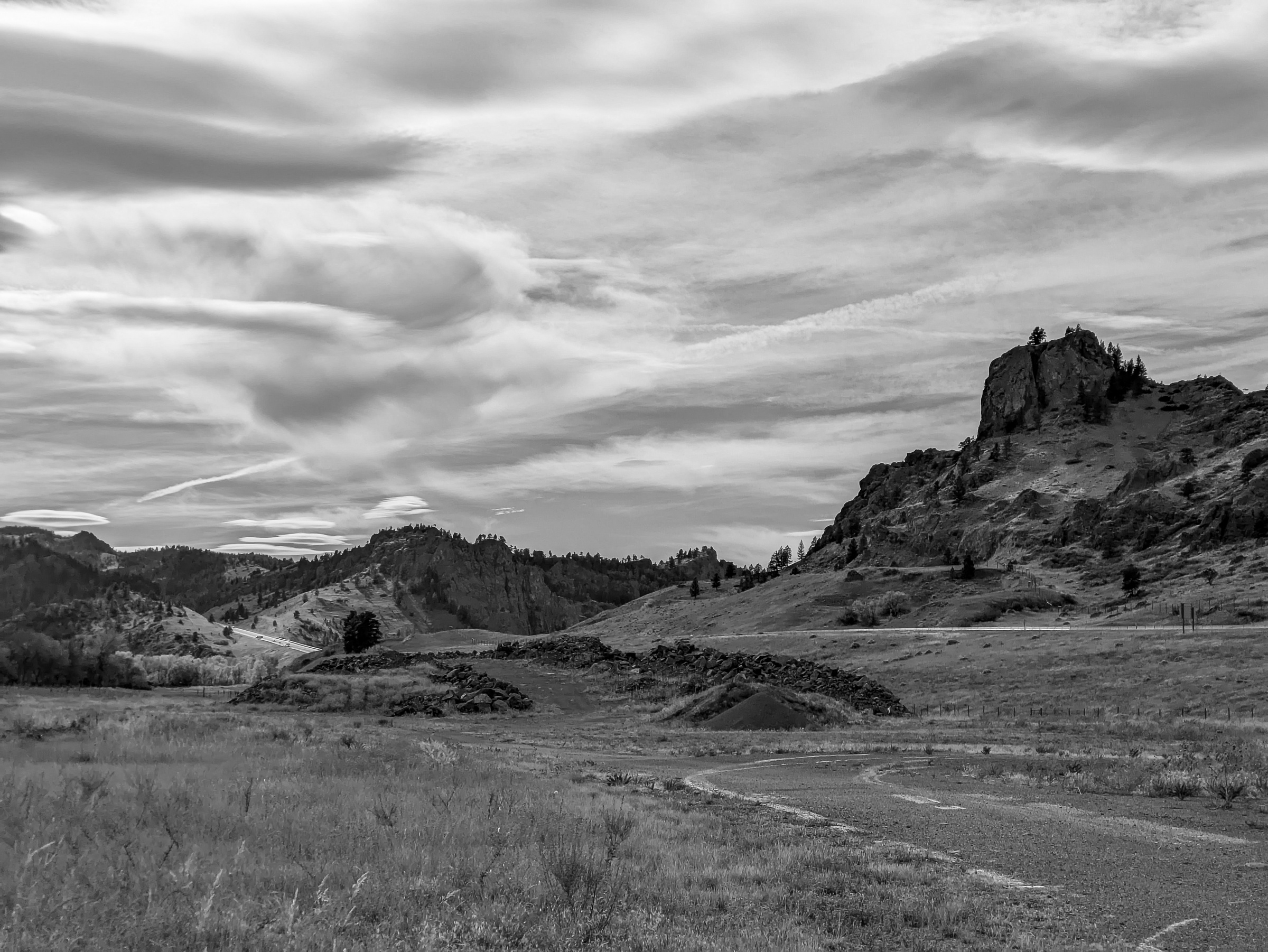 A rocky peak rises above rolling hills under a dramatic cloudy sky in black and white.