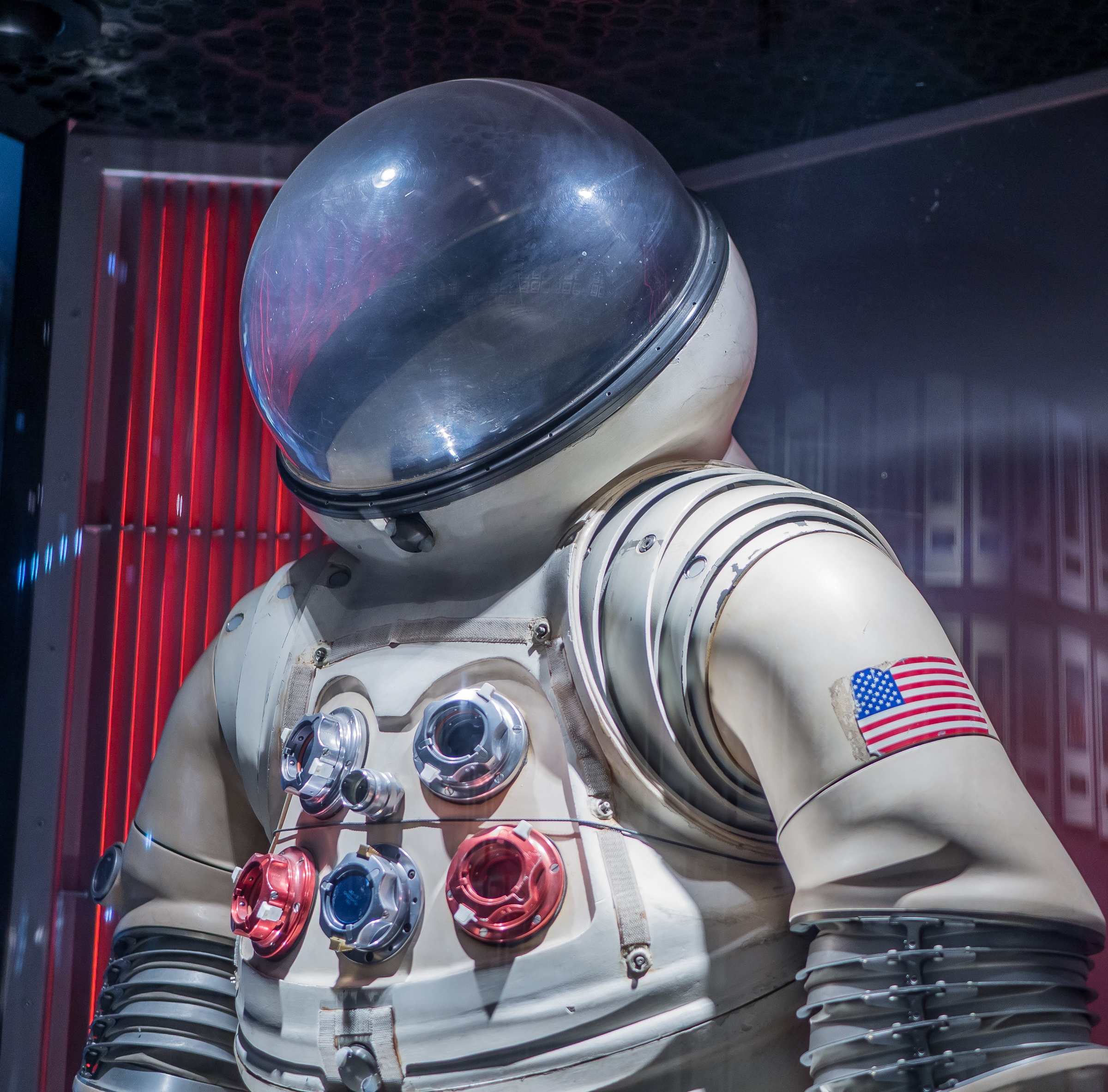 A white spacesuit with an American flag patch is displayed against a red and blue textured wall.