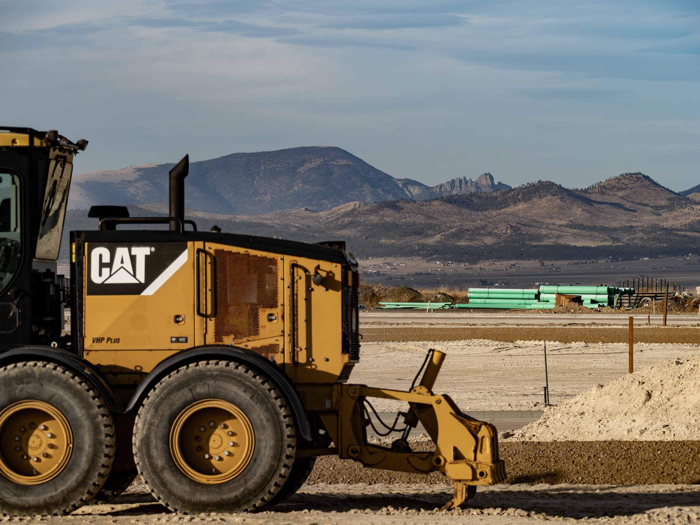 A yellow Caterpillar grader sits in a construction site with mountains visible in the distance.