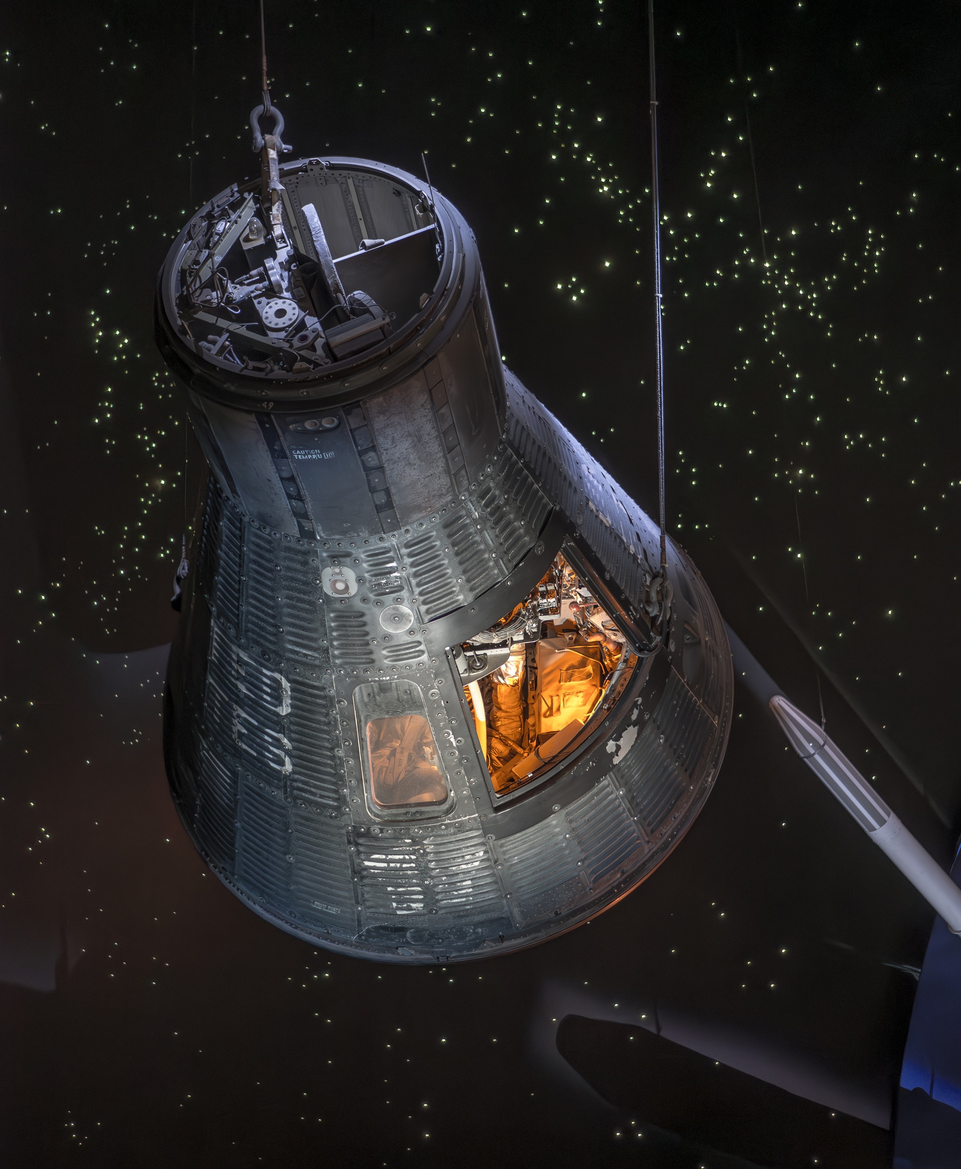 A blackened Apollo command module hangs suspended against a starry, dark background.