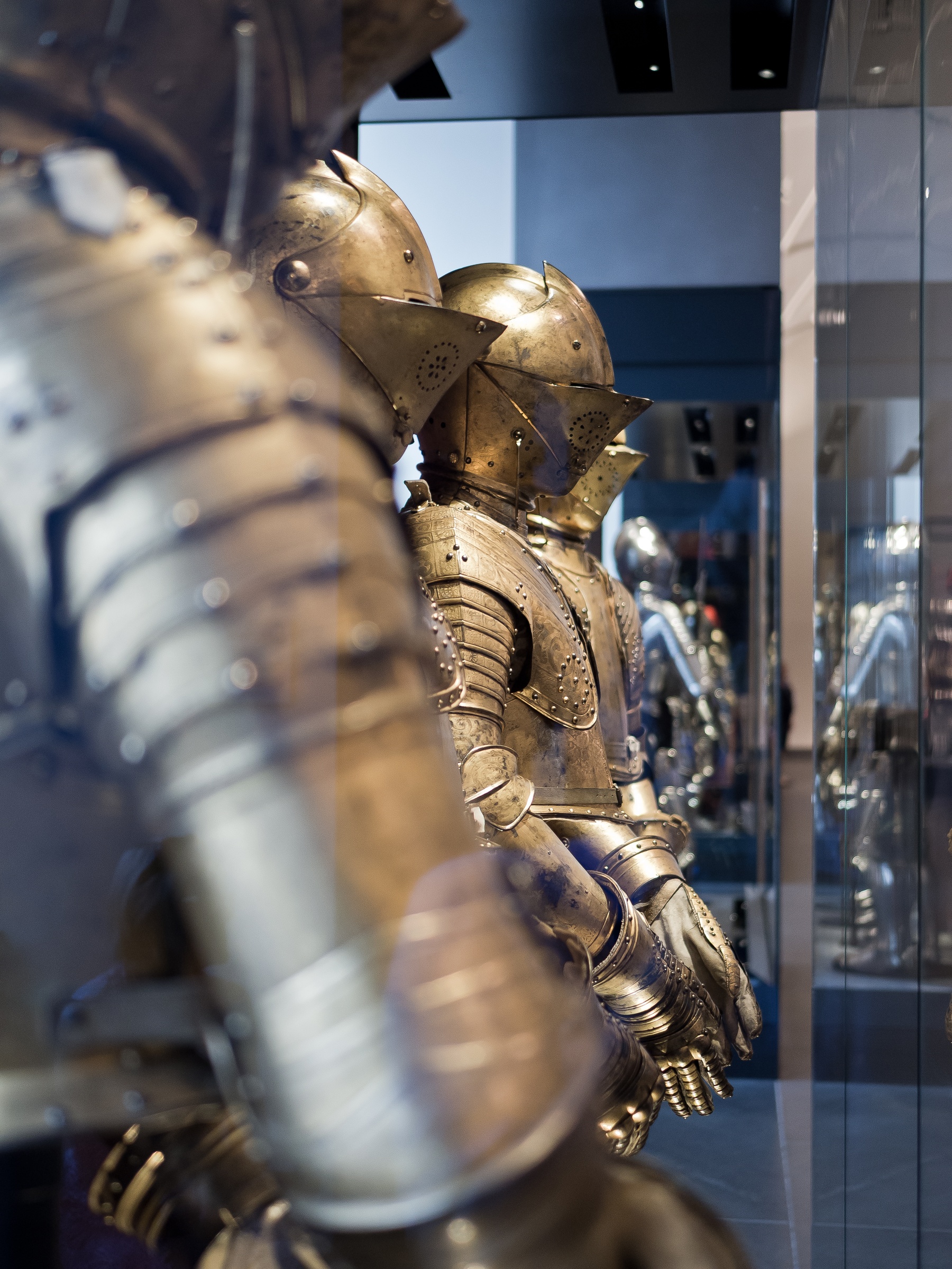Two antique suits of polished metal armor are displayed behind glass in a museum.