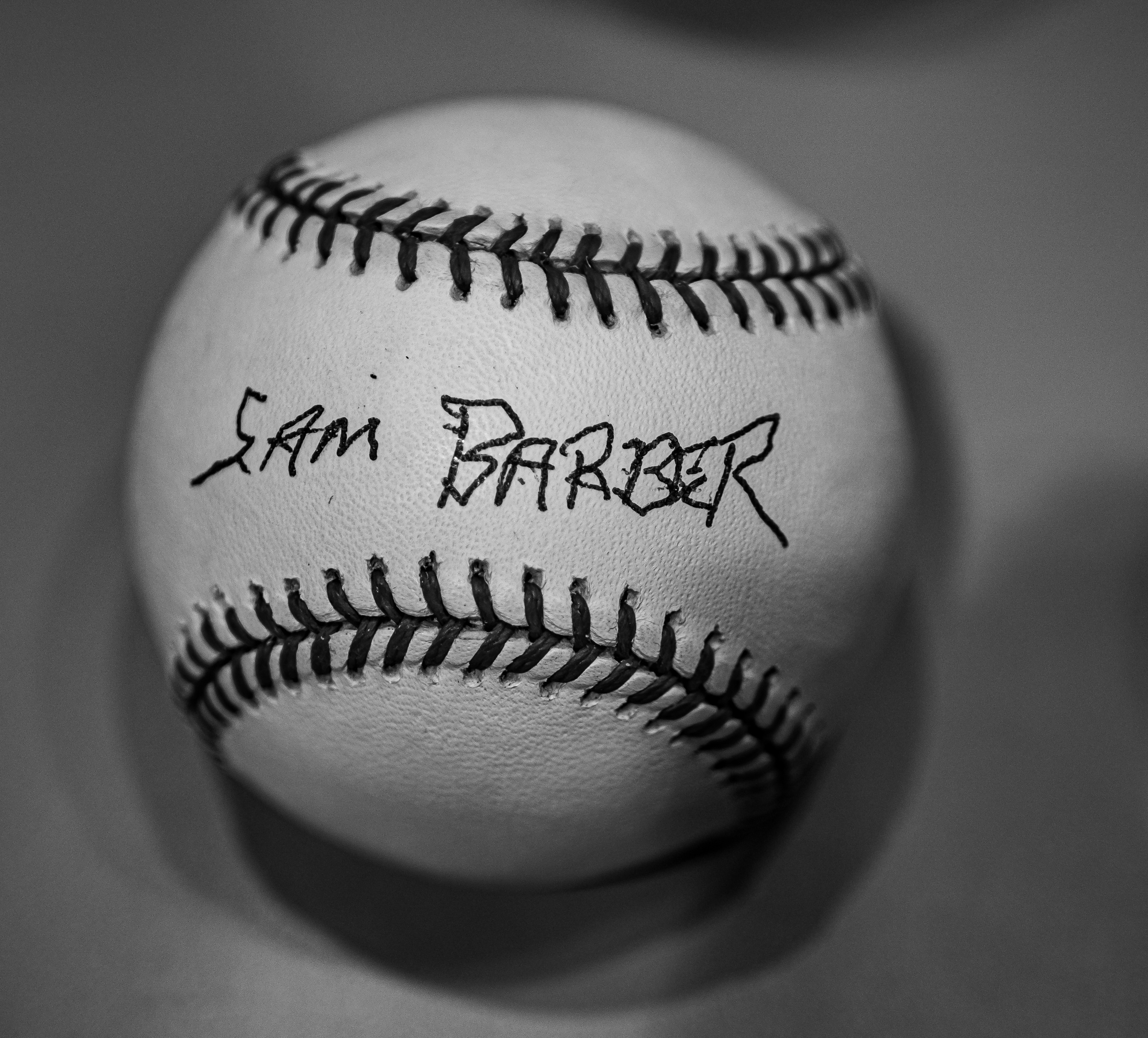 A baseball with the name "Sam Barber" scrawled on it rests against a gray surface.