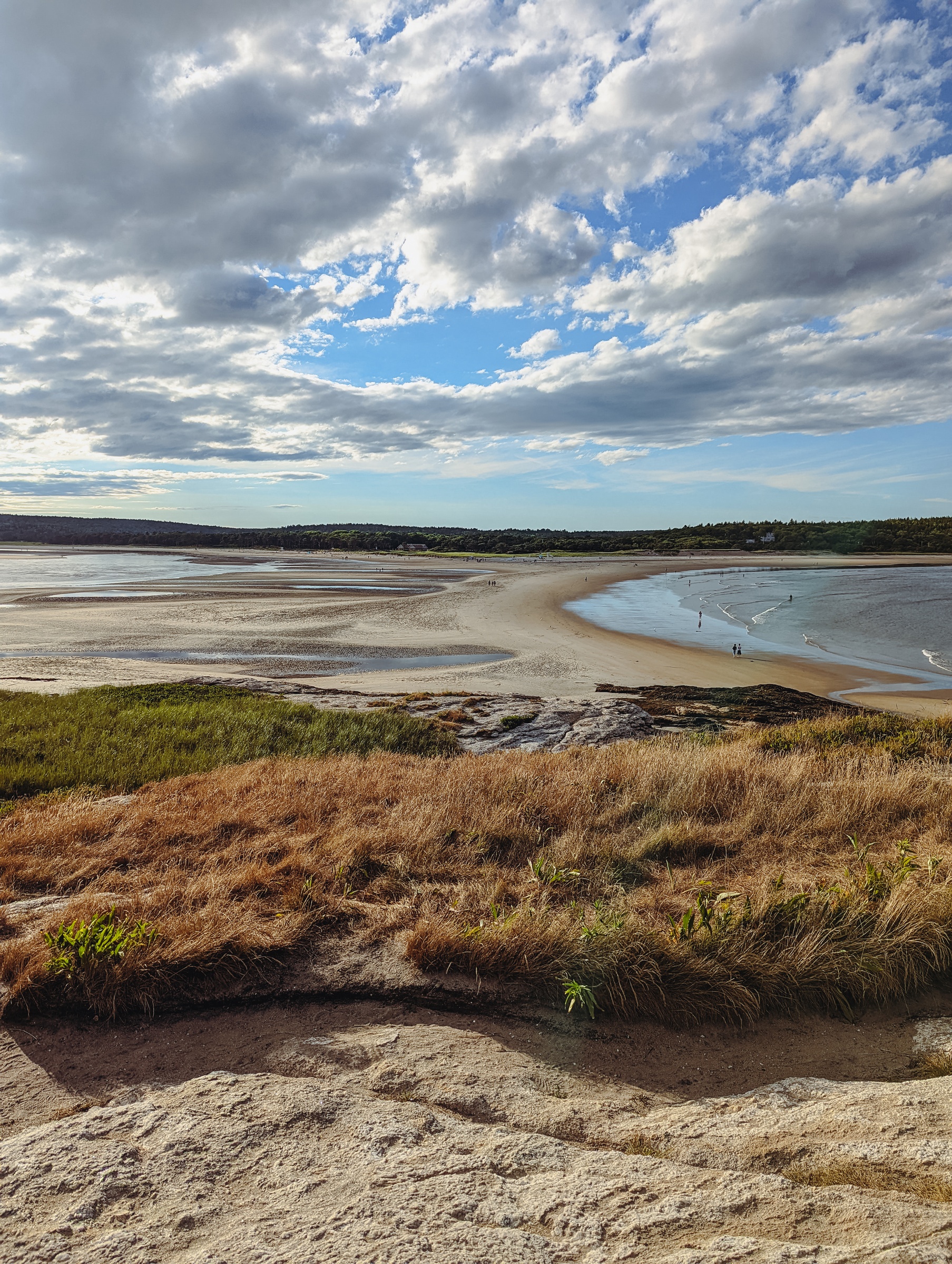Grassy cliffs overlook a wide sandy beach and water under a cloudy blue sky.