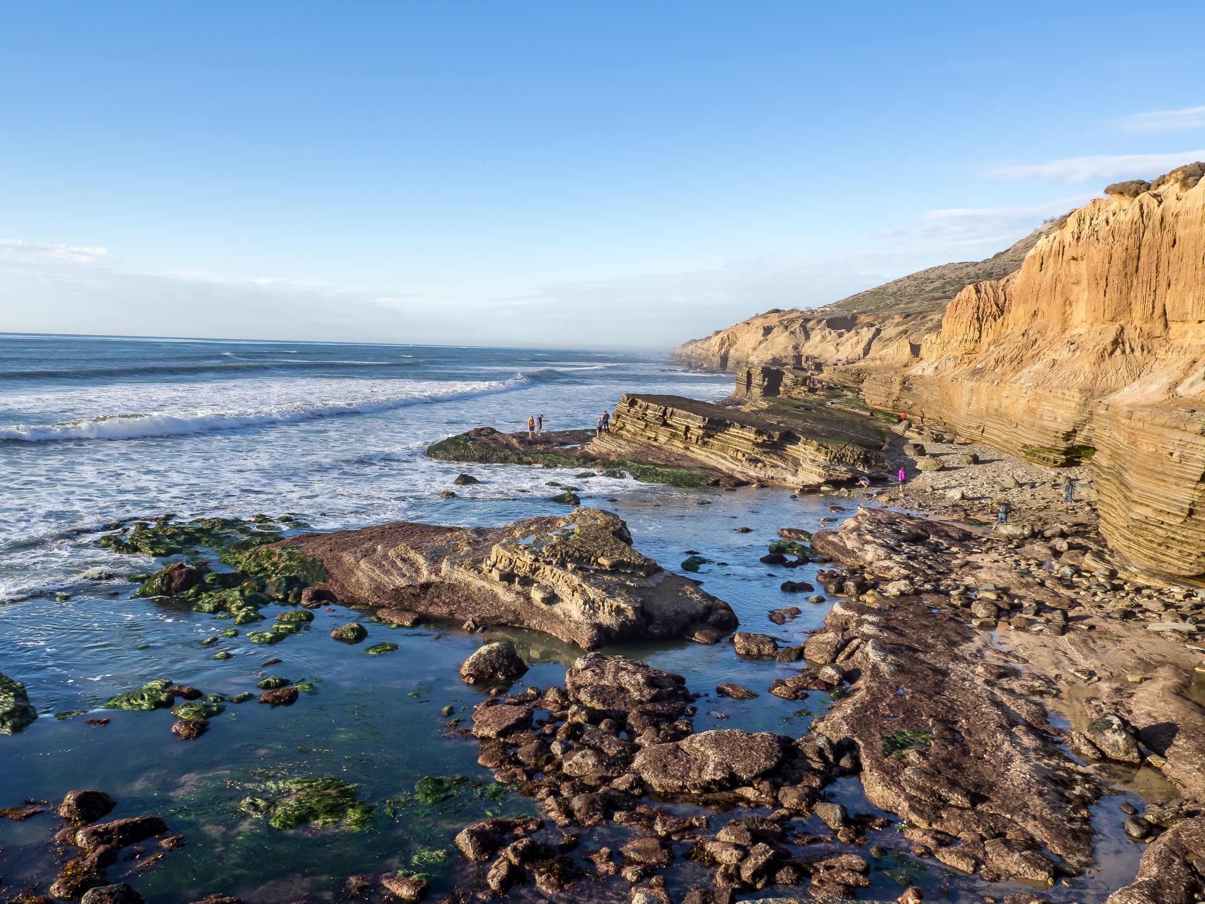 Ocean waves roll onto a rocky shoreline beneath layered sandstone cliffs and a clear blue sky.