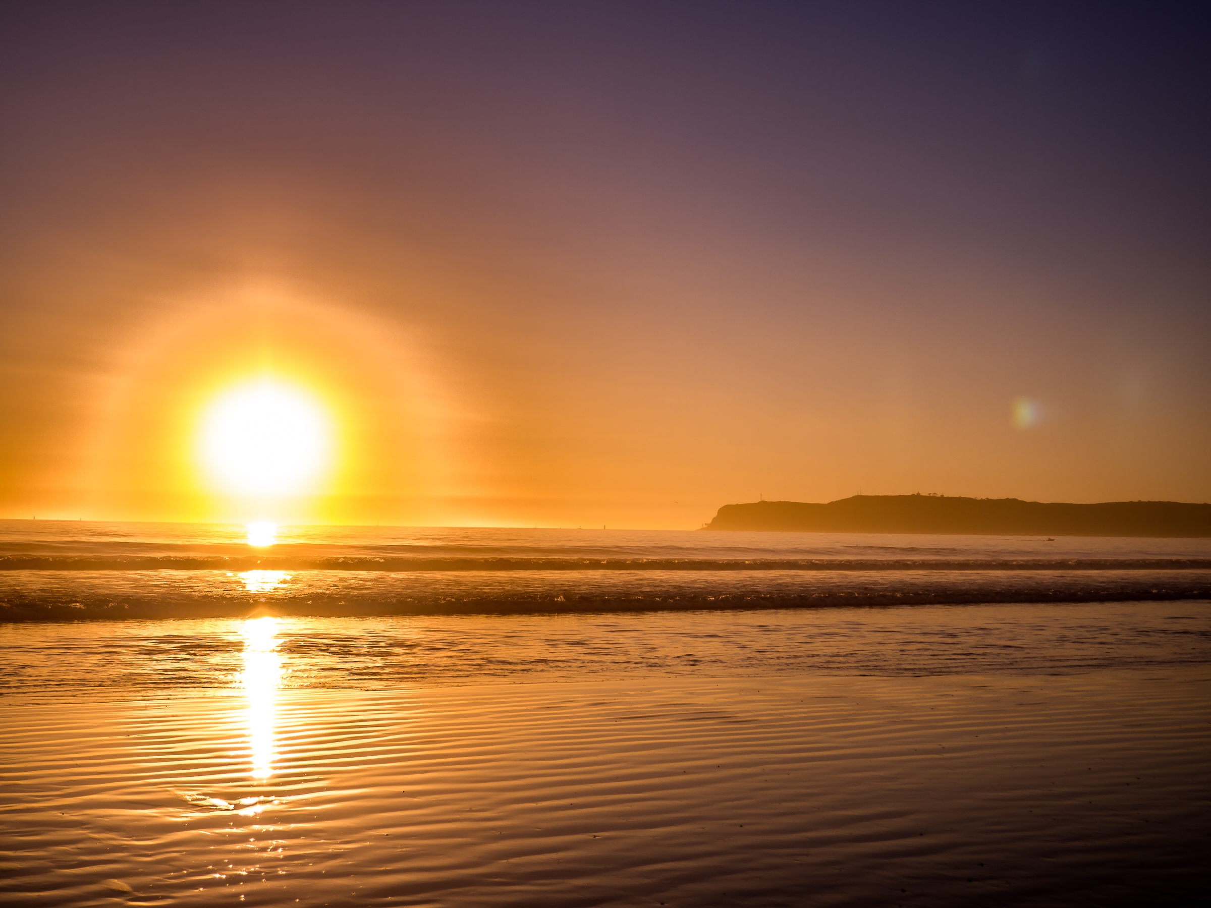 The sun sets over a sandy beach, casting reflections on the wet sand near a distant coastline.