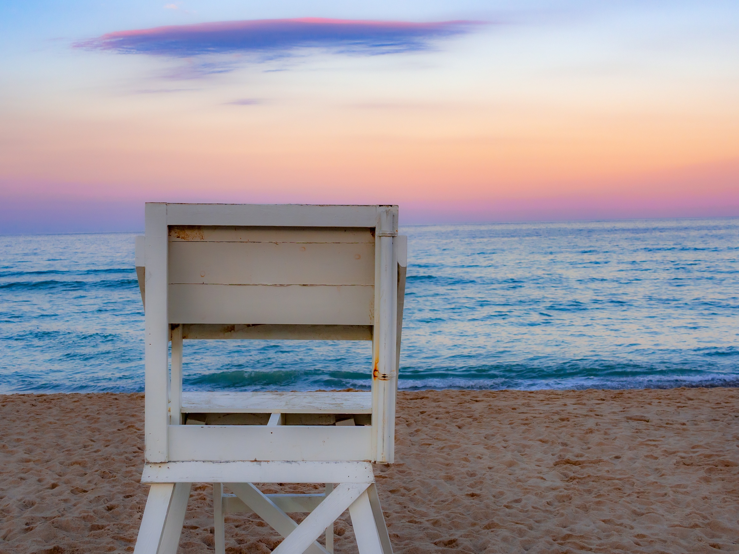 A white lifeguard chair stands on a sandy beach facing the ocean under a colorful sky.