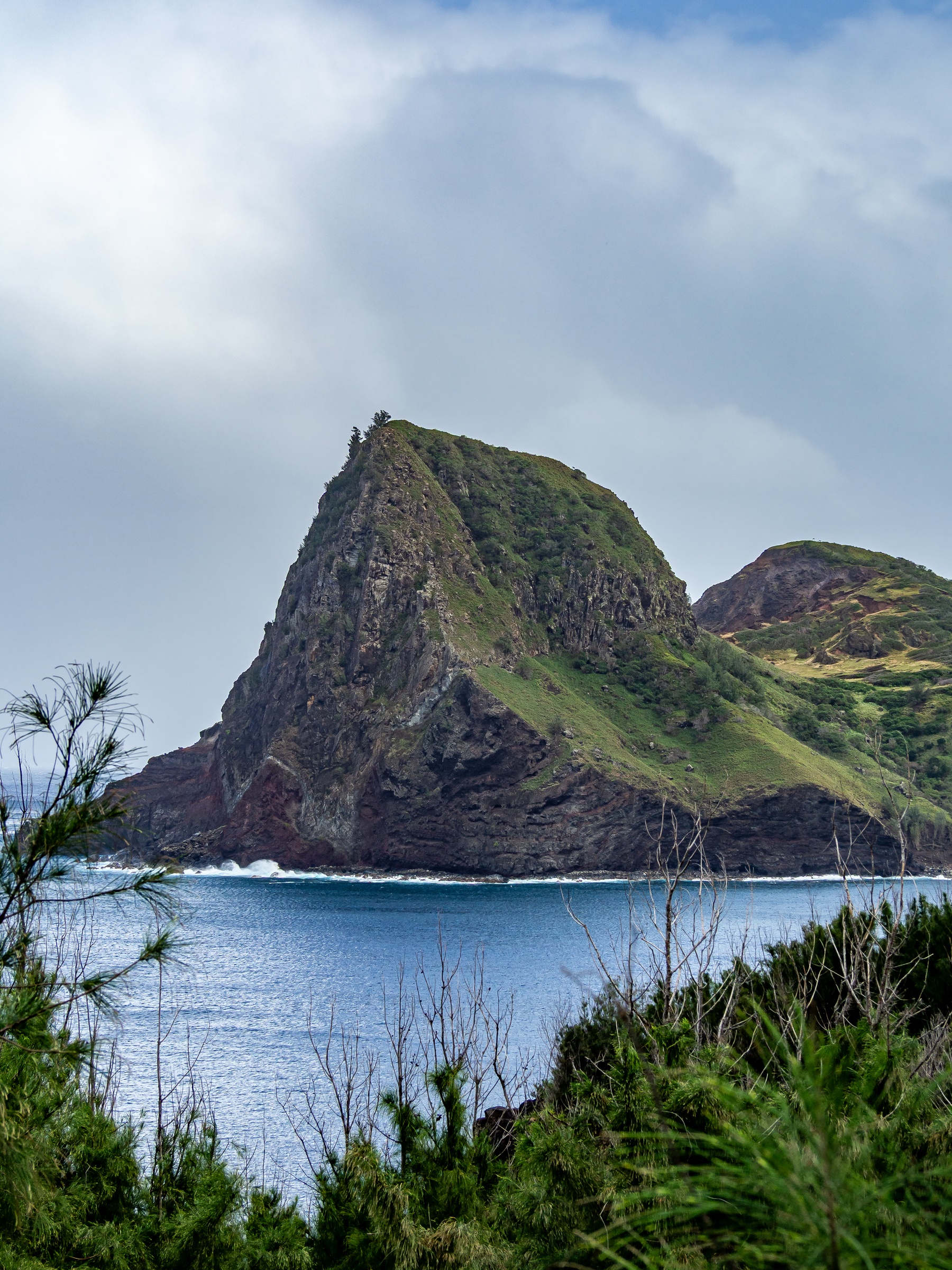 A steep, green rocky cliff rises from a blue ocean under a cloudy sky.