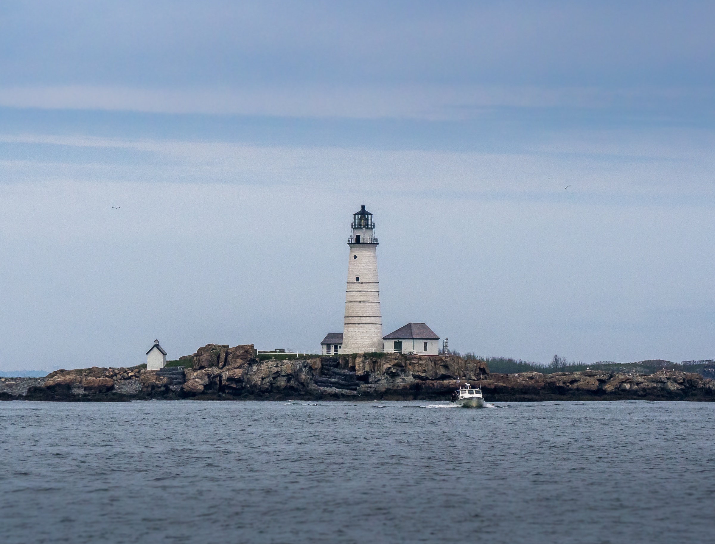 A white lighthouse and small buildings stand on a rocky island surrounded by water.