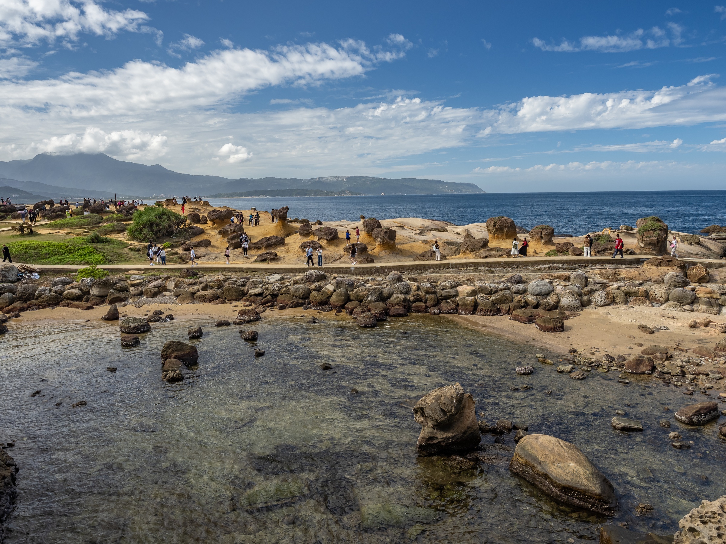 People walk along a rocky shoreline with clear shallow water and distant mountains under a blue sky.