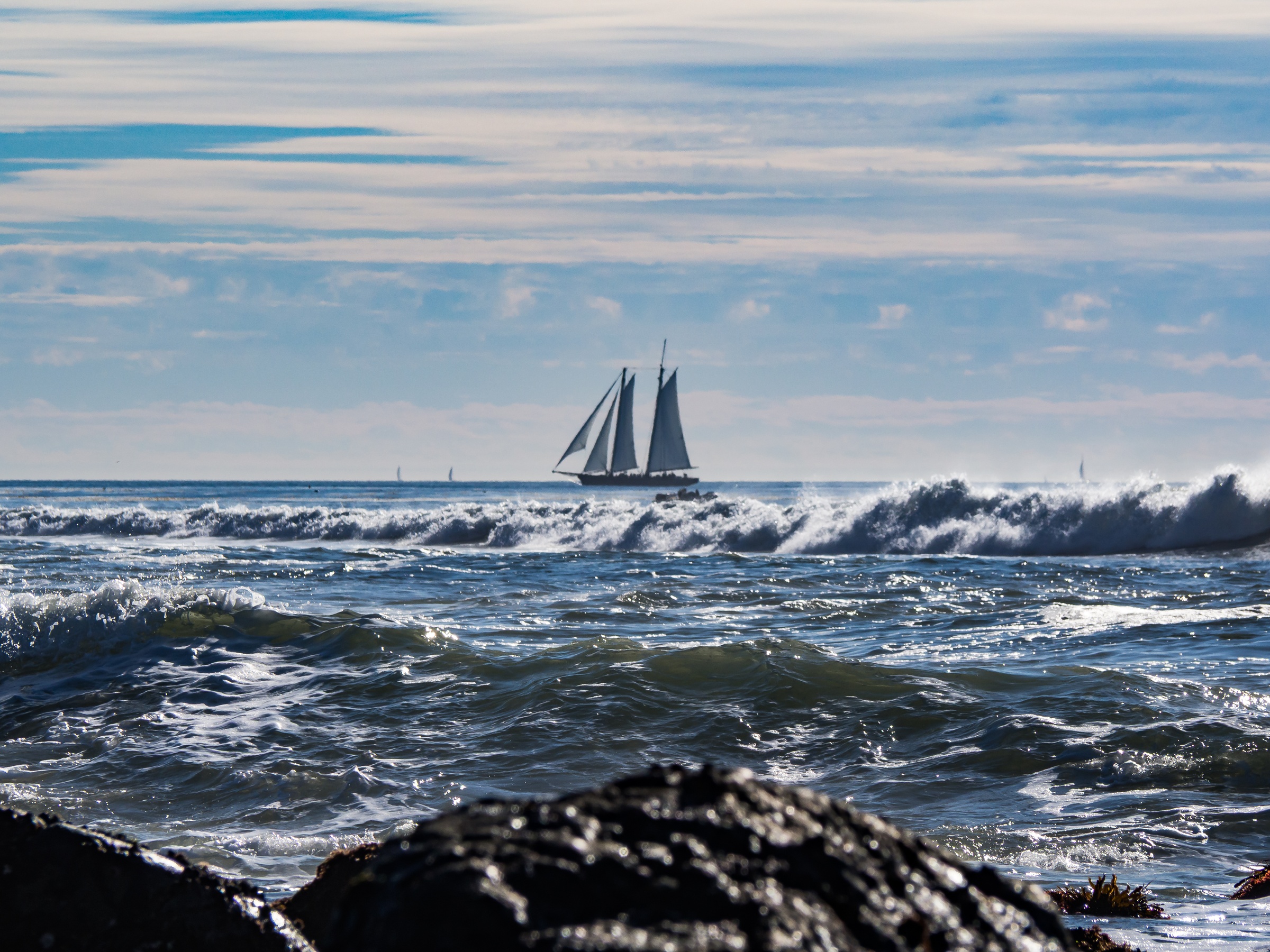 A two-masted sailboat sails on an ocean with crashing waves and dark rocks.