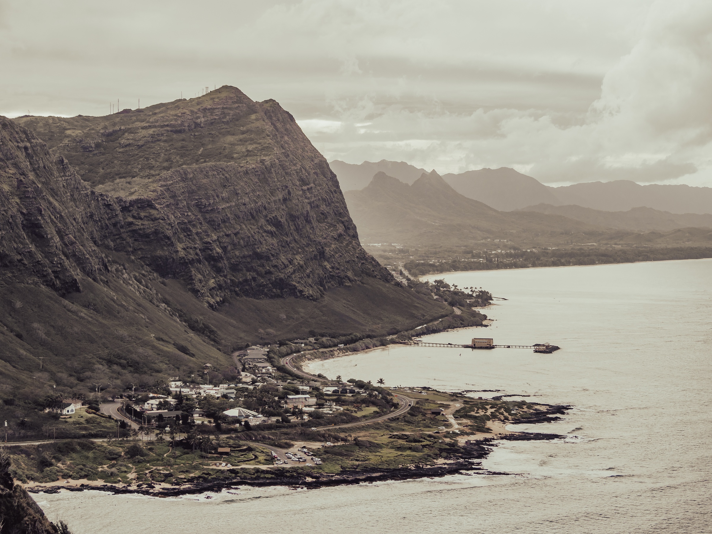 A massive rocky cliff towers over a small coastal town and pier beside the ocean.